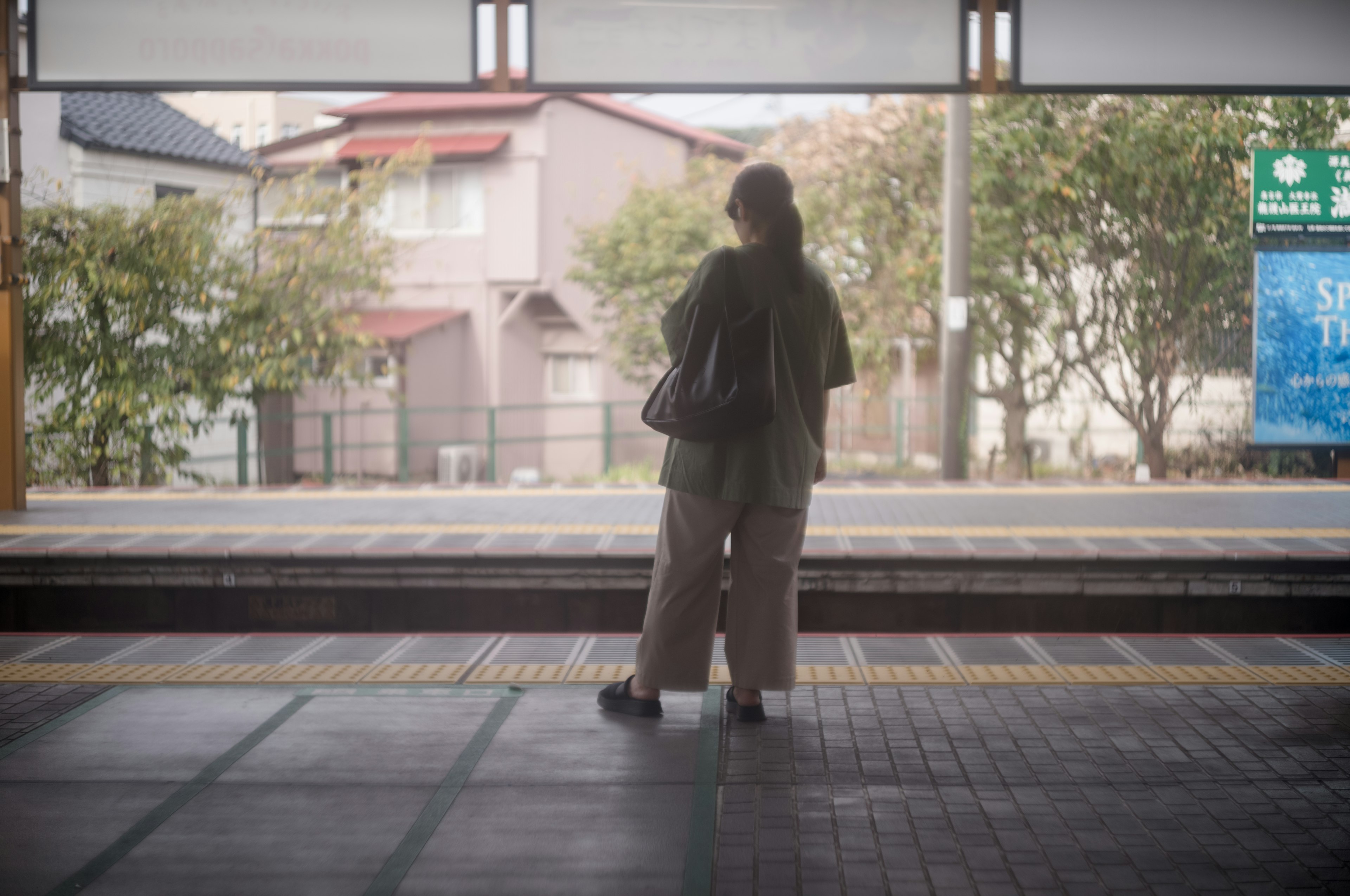 駅のプラットフォームで待つ女性の後ろ姿周囲に木々と住宅