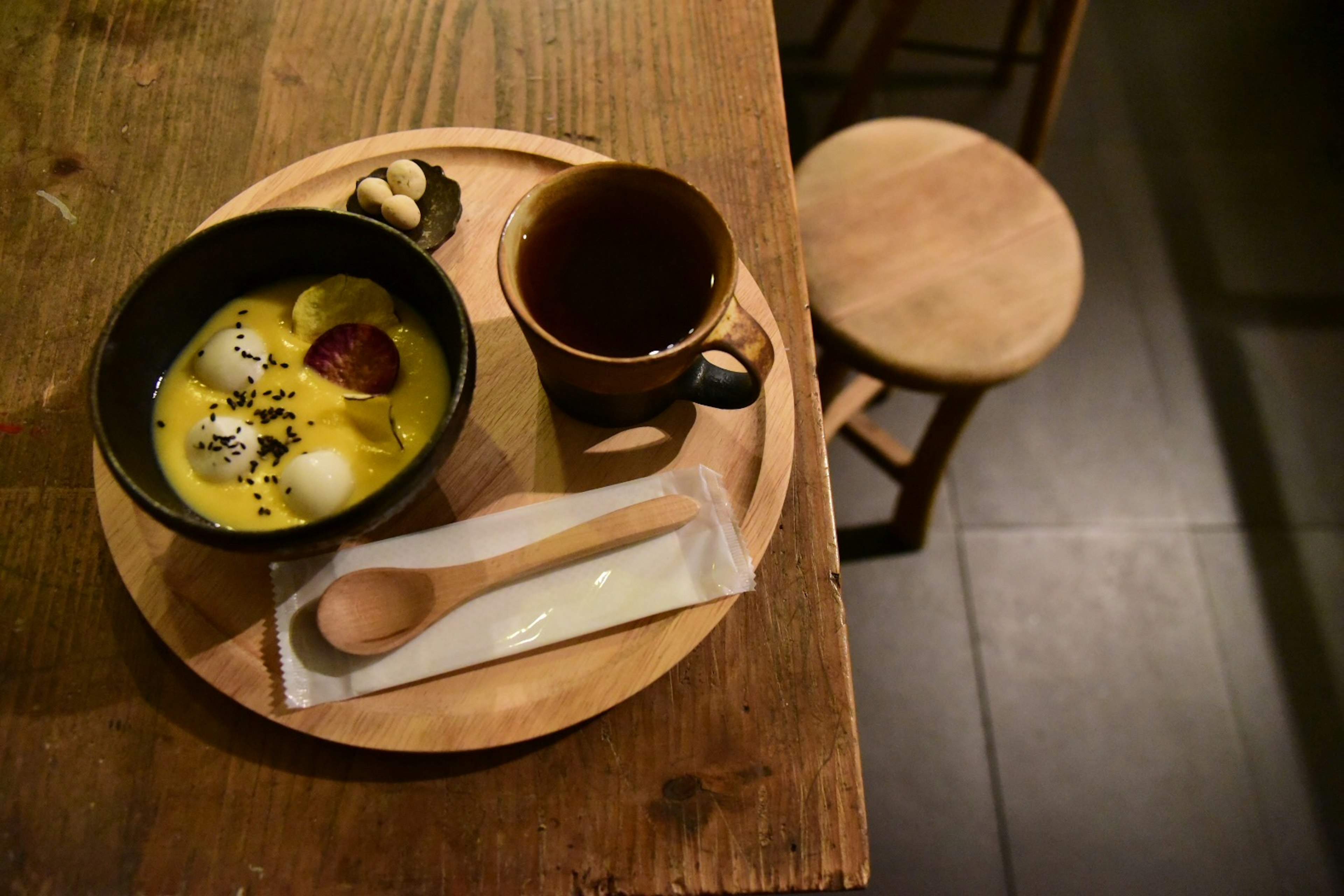 A wooden tray with a black bowl of dessert and mochi balls alongside a cup of coffee