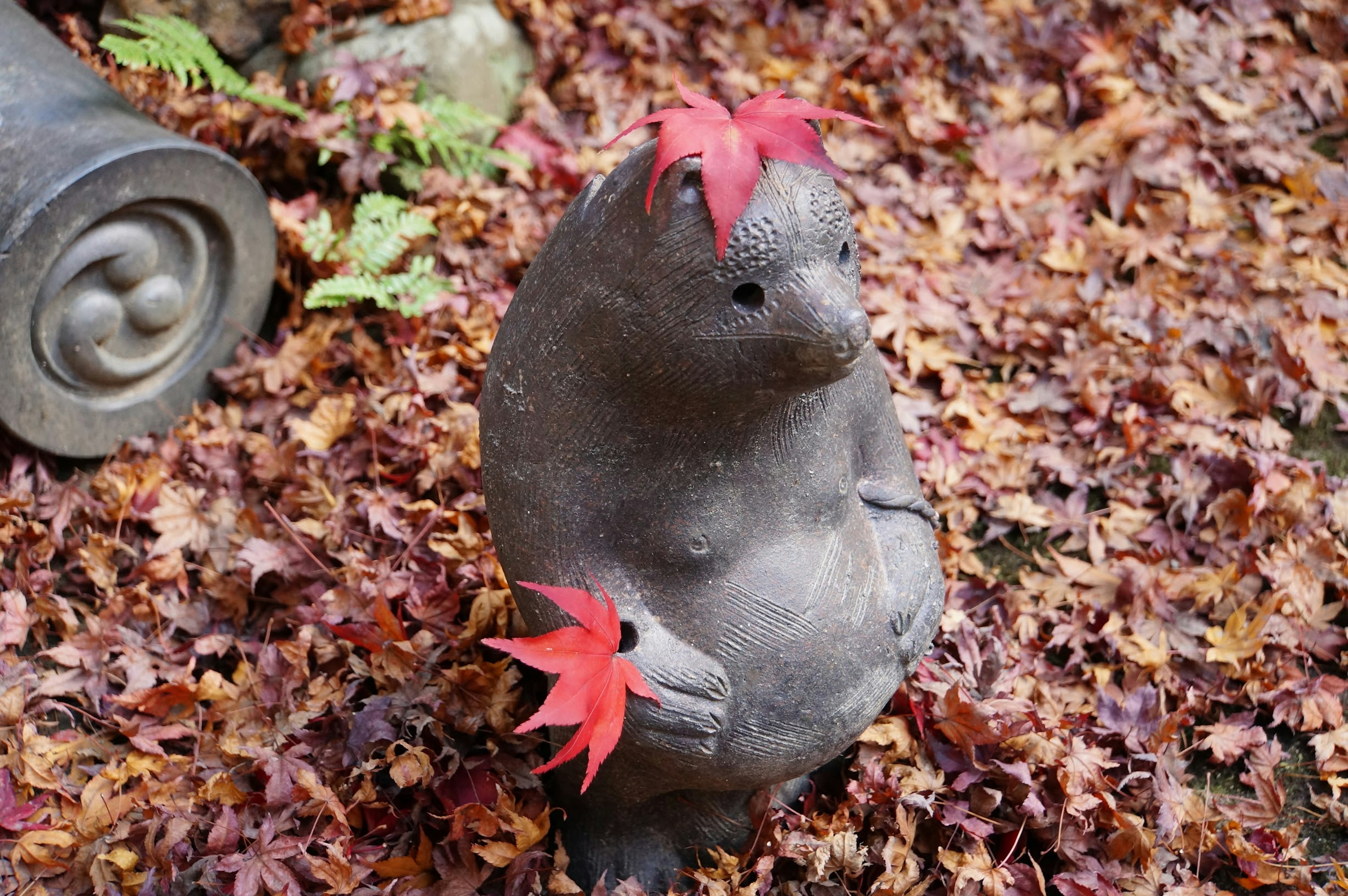 赤い葉を持つ石のクマの彫刻が落ち葉の中に立っている