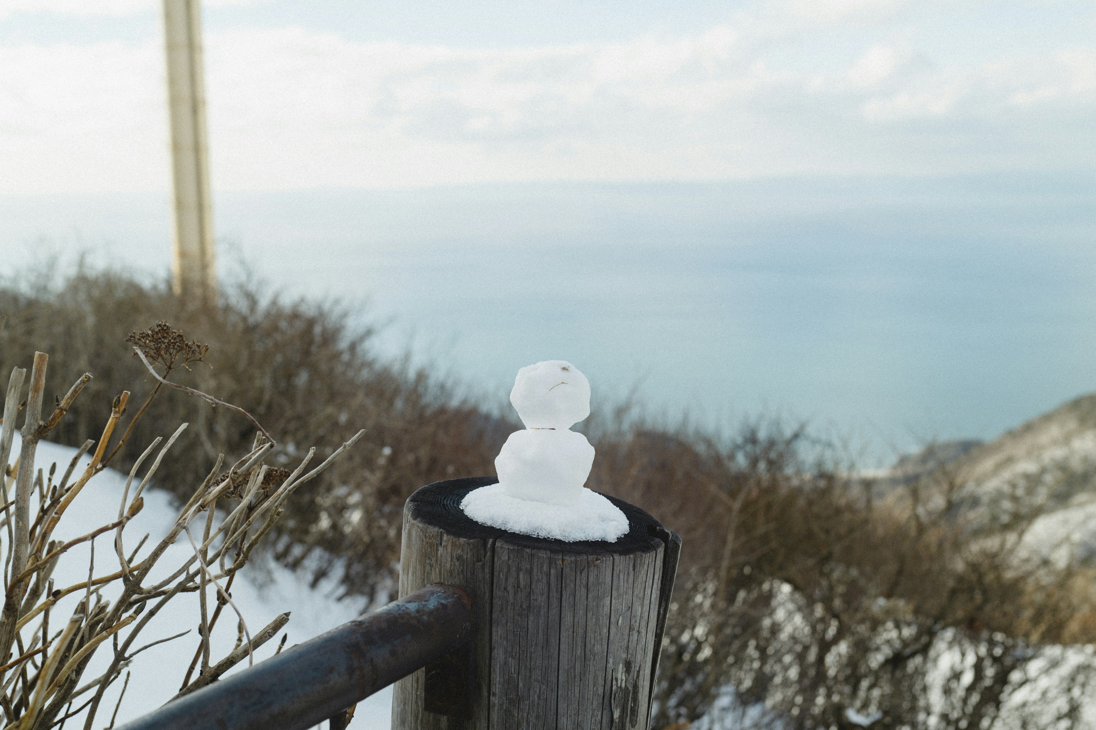 A small snowman made of snow sitting on top of a wooden post with a view of the sea