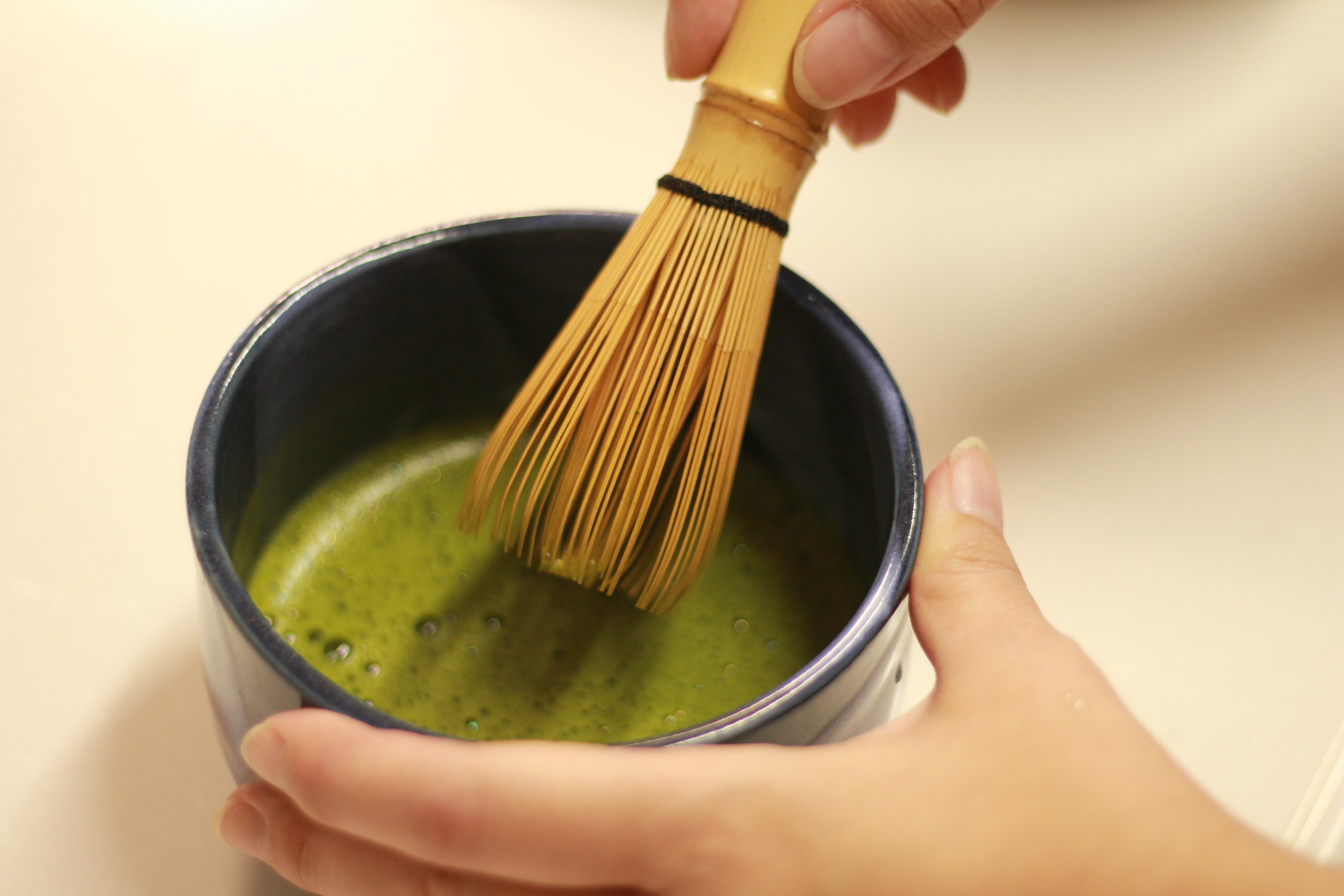 Hand holding a bamboo whisk in a blue bowl of matcha tea