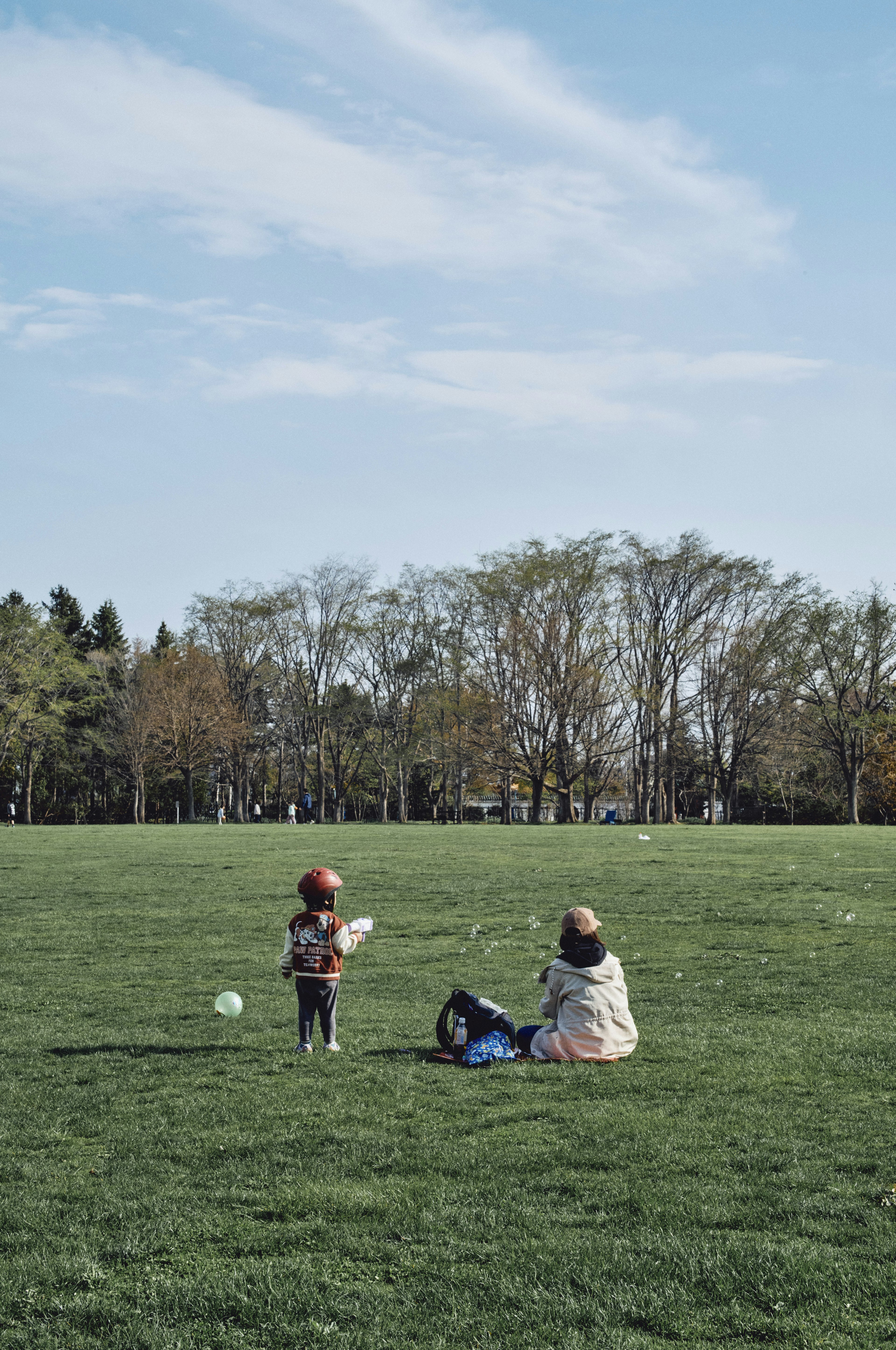 A child playing in a green park with an adult sitting on a blanket