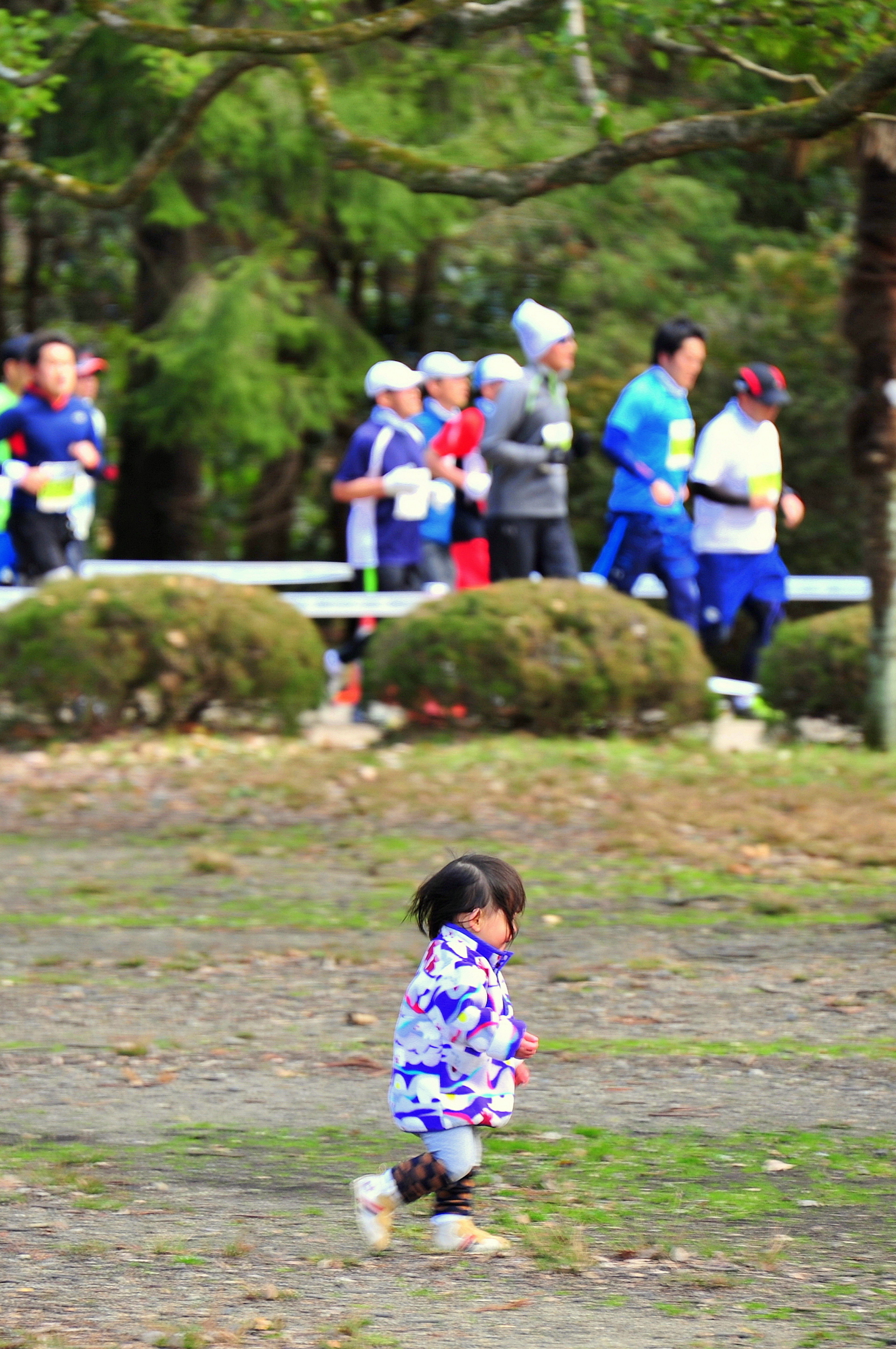 A young child wearing a blue coat running in a park with runners in the background