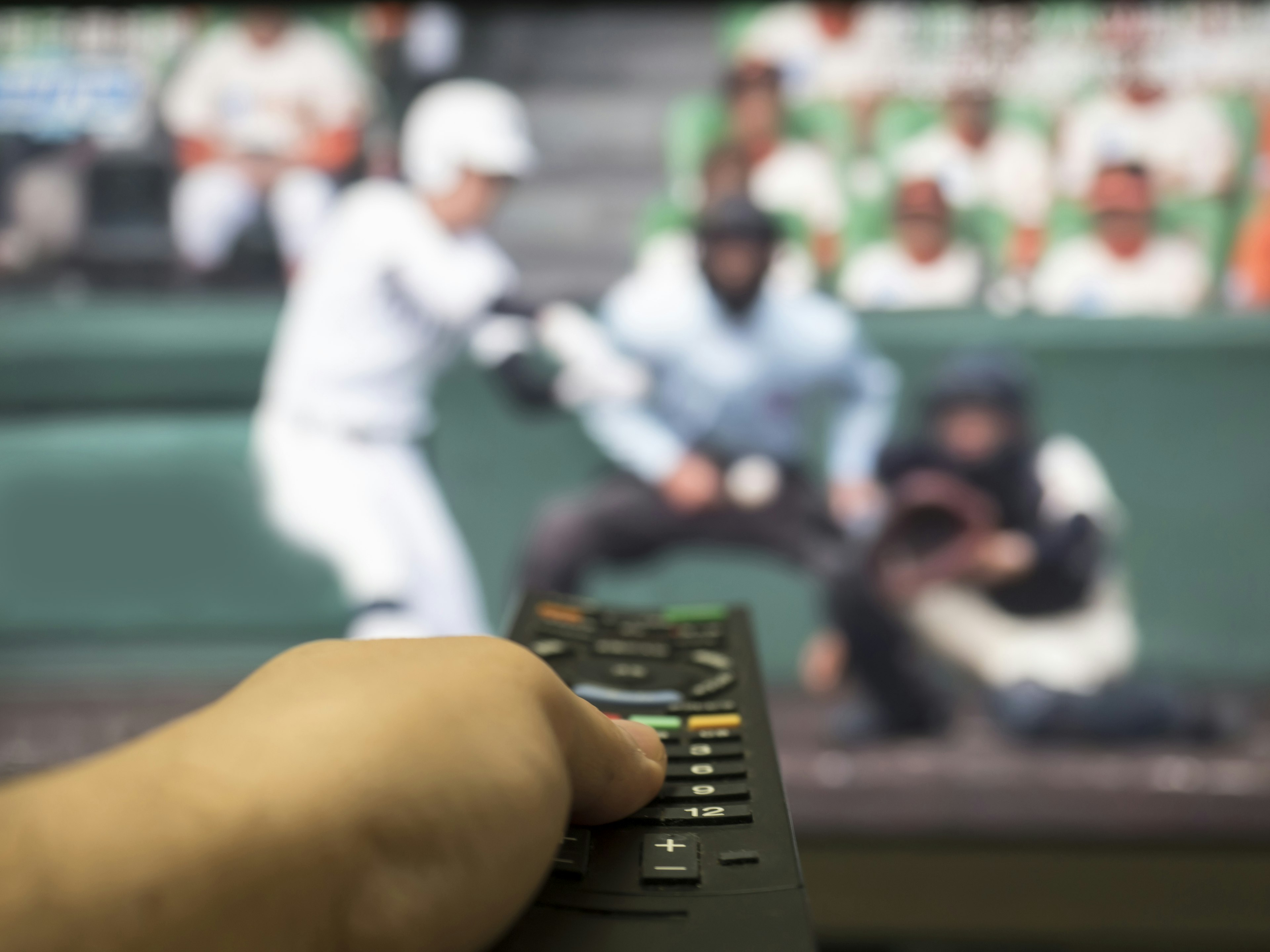 Hand, die eine Fernbedienung hält, mit einem Baseballspiel auf dem Fernseher