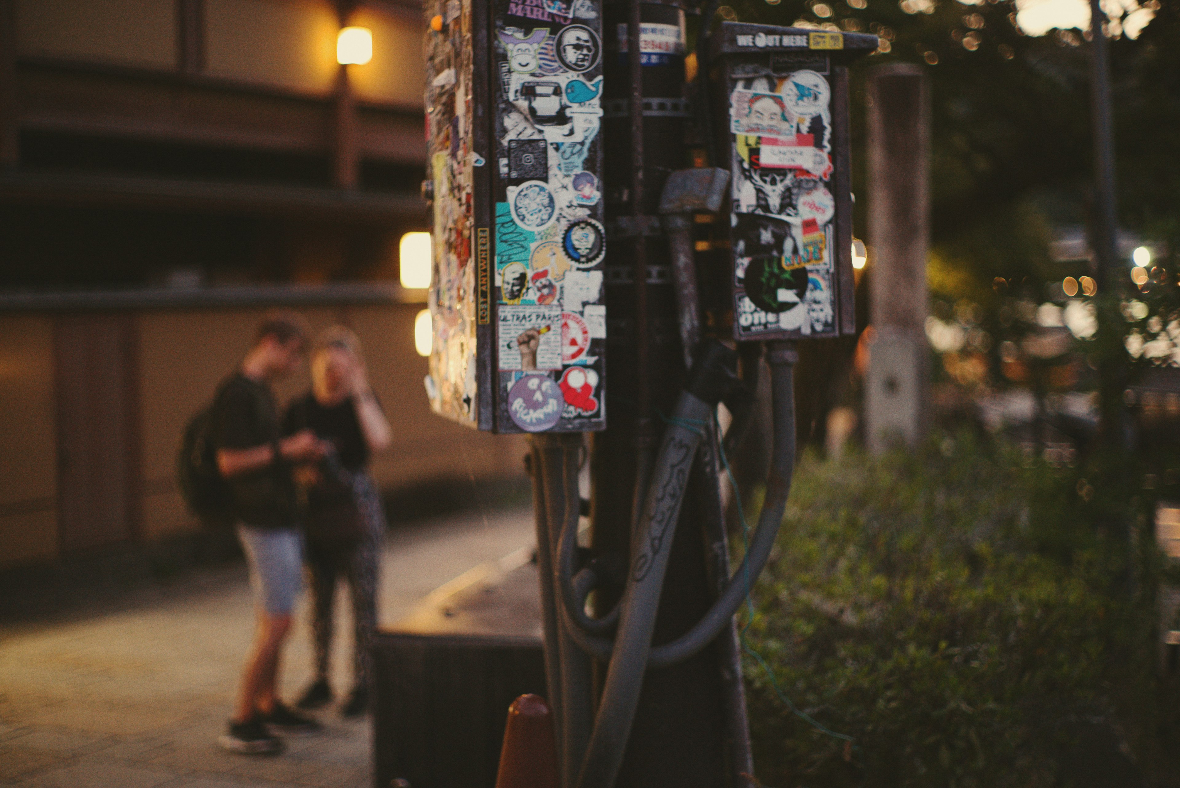 A public phone covered in stickers at night with two people standing nearby