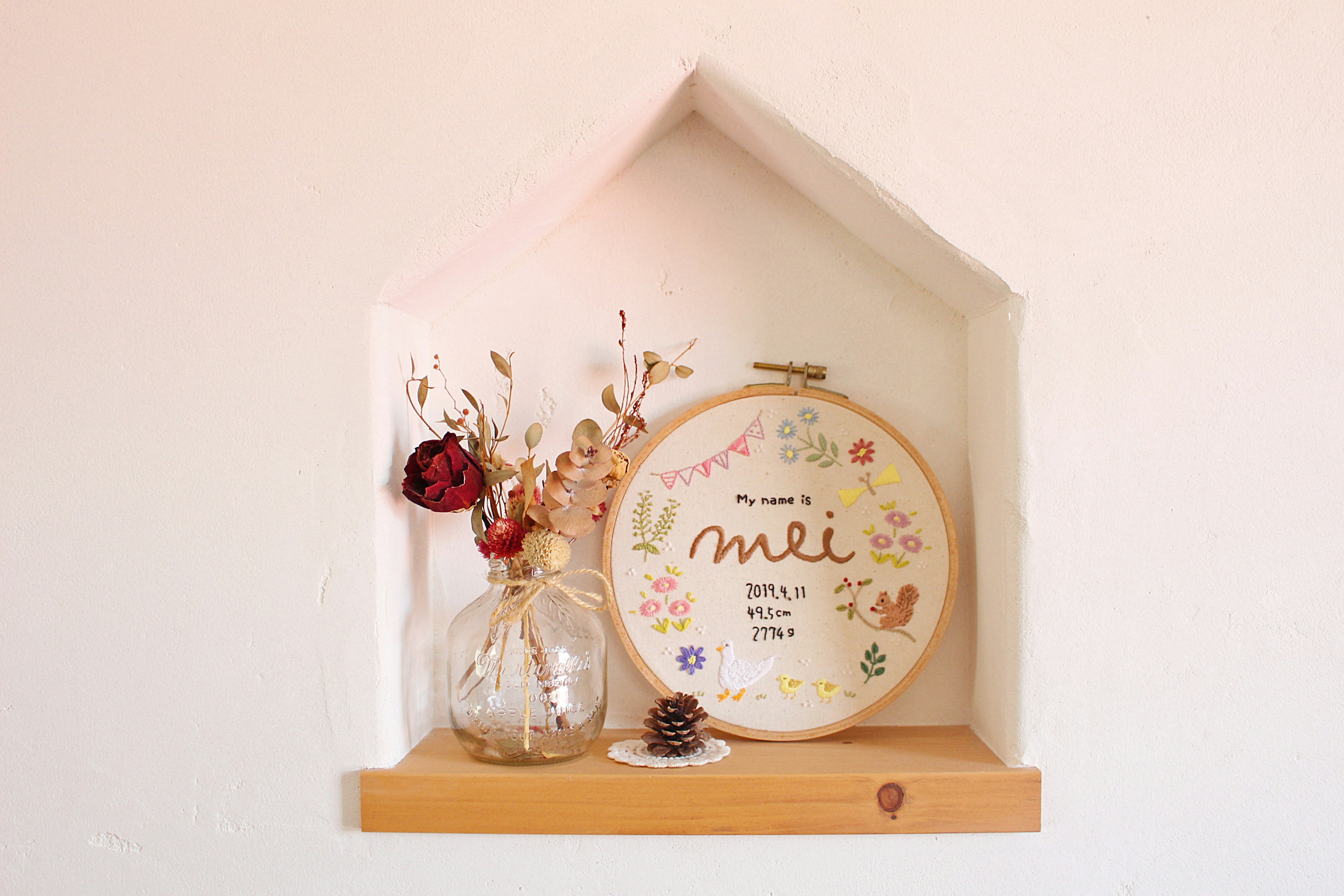 A small house-shaped shelf on a wall displaying an embroidered frame and a vase