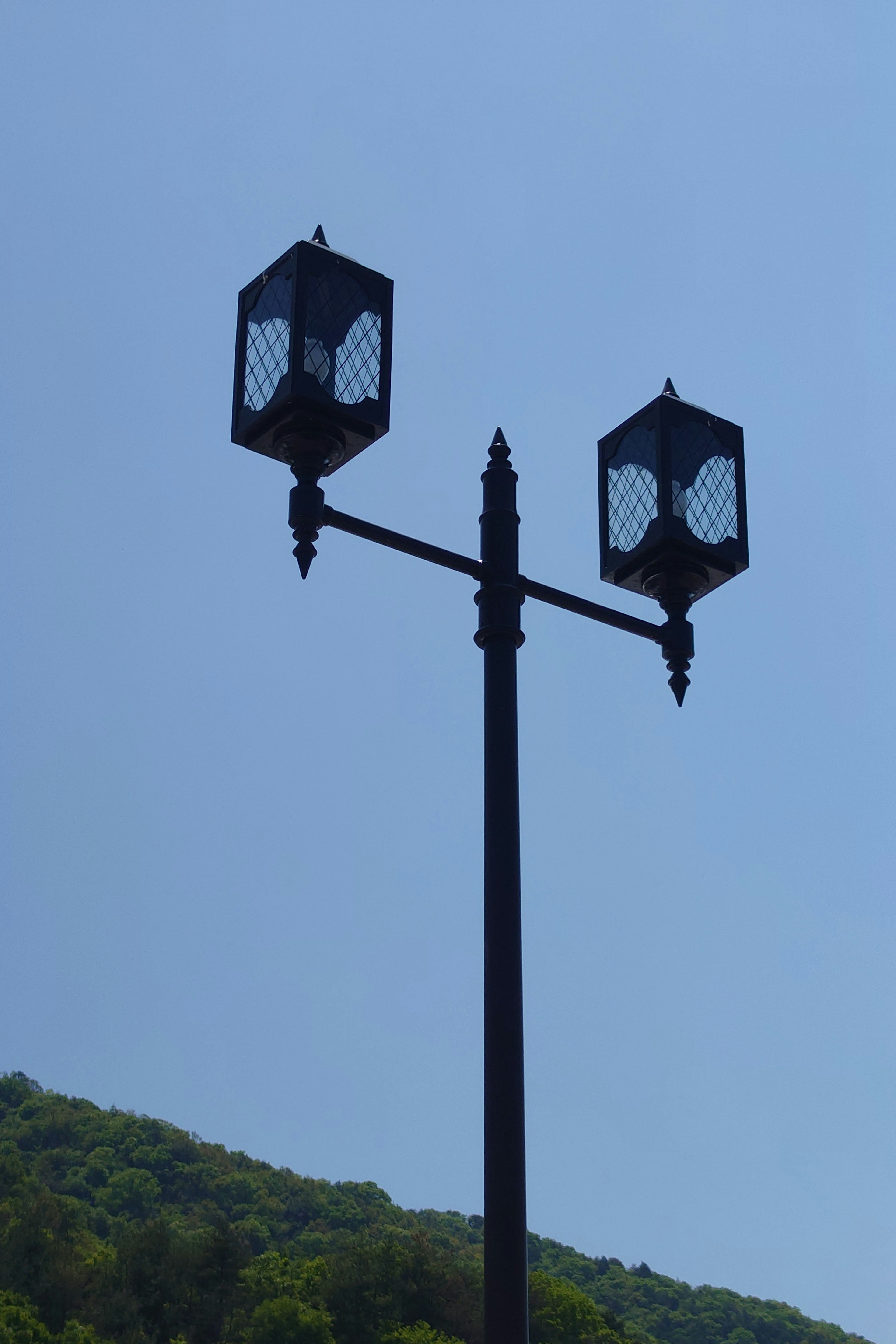 Farola con dos linternas bajo un cielo azul y una colina verde