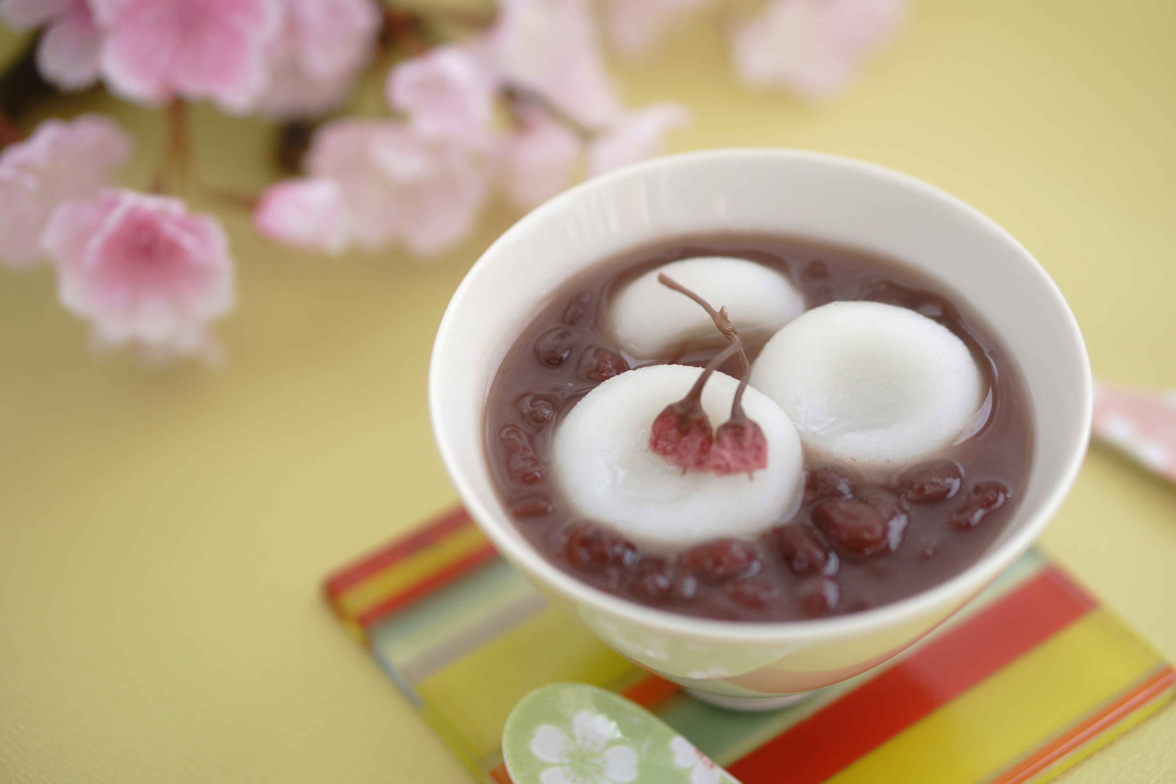 Bowl of sweet red bean soup with three white rice dumplings and a cherry on top surrounded by pink cherry blossoms