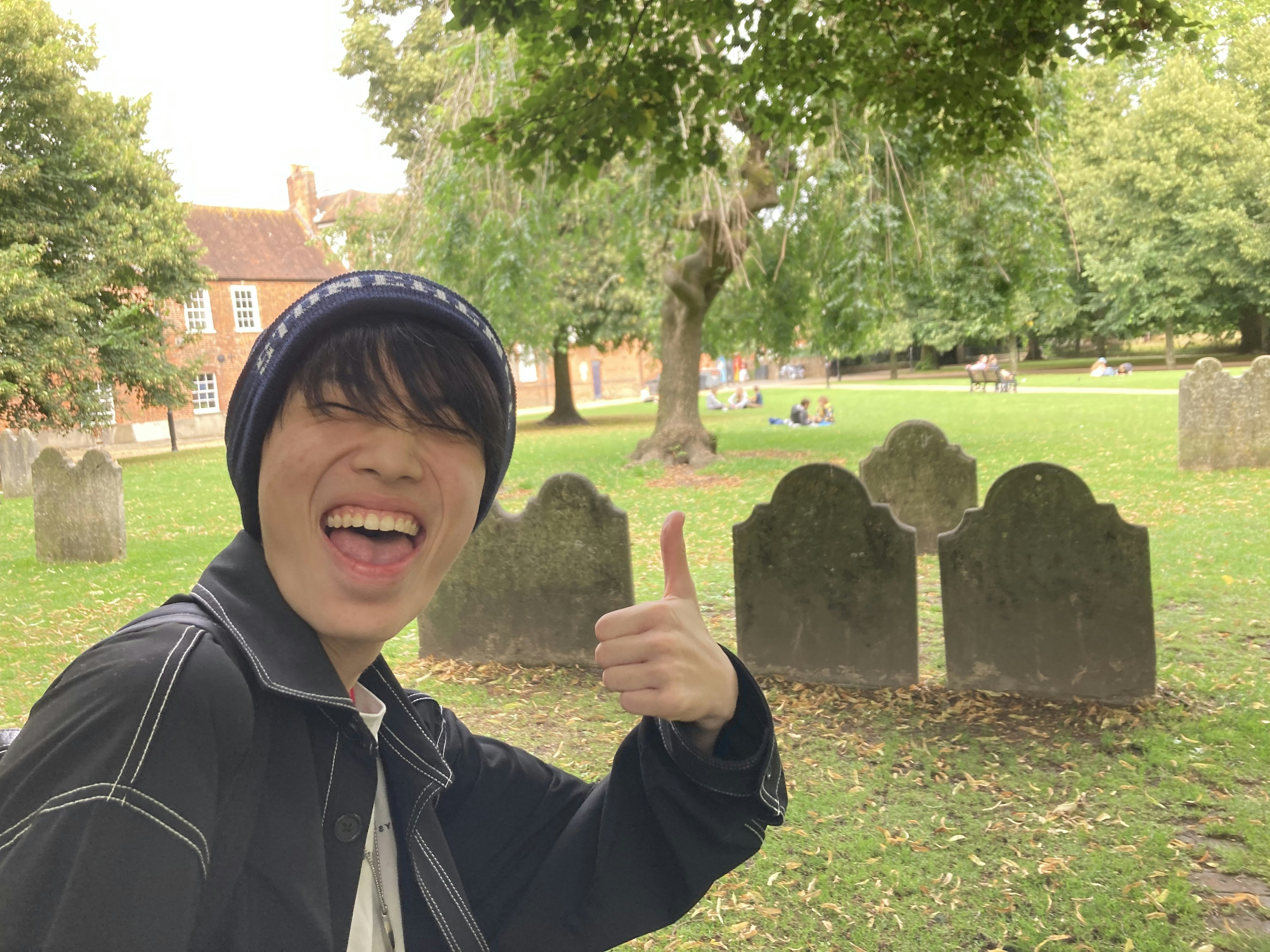 A young man smiling and giving a thumbs up in a park with gravestones