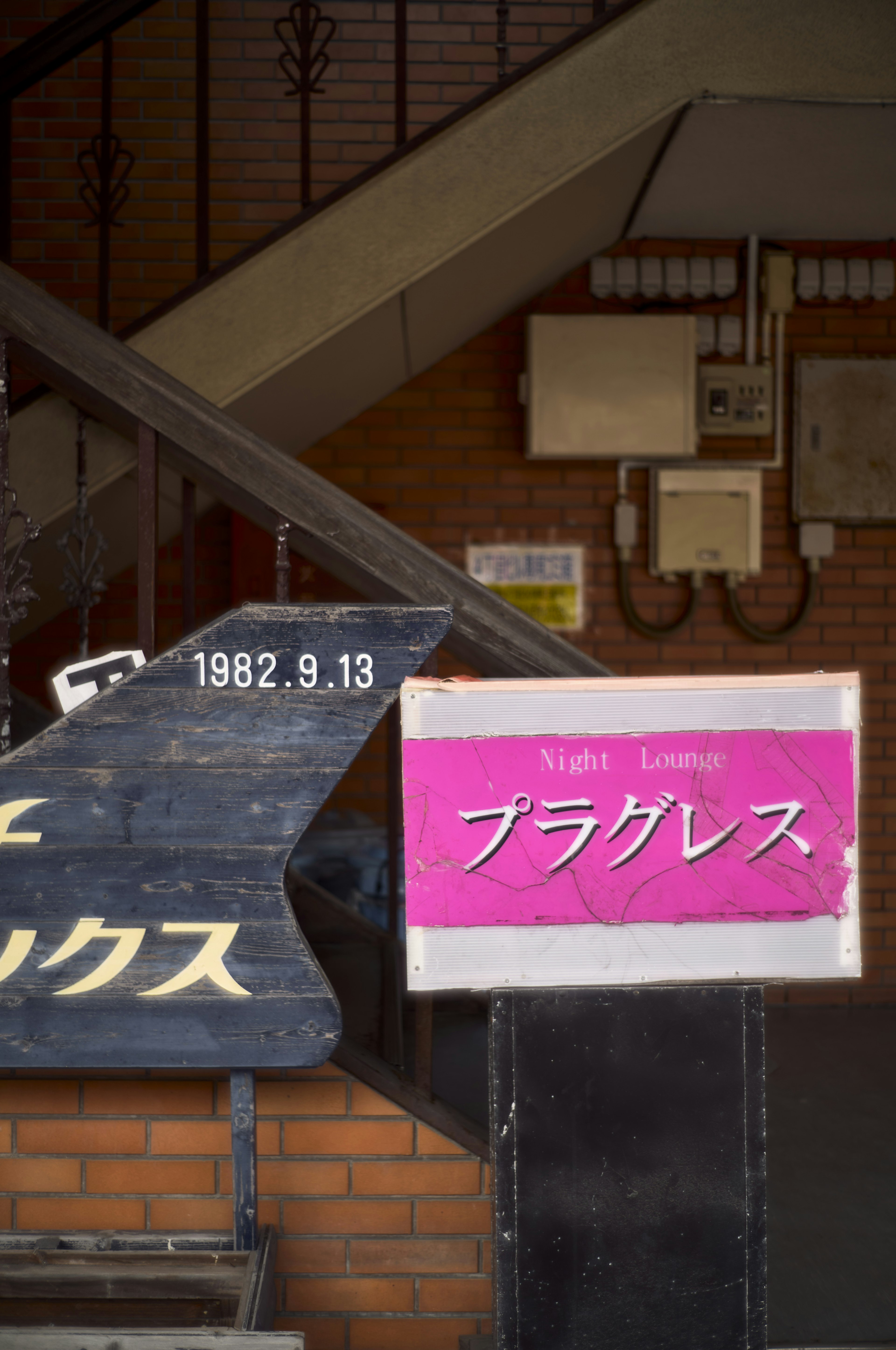 Ein rosa Schild mit der Aufschrift 'プラグレス' in der Nähe einer Treppe mit Ziegelwand