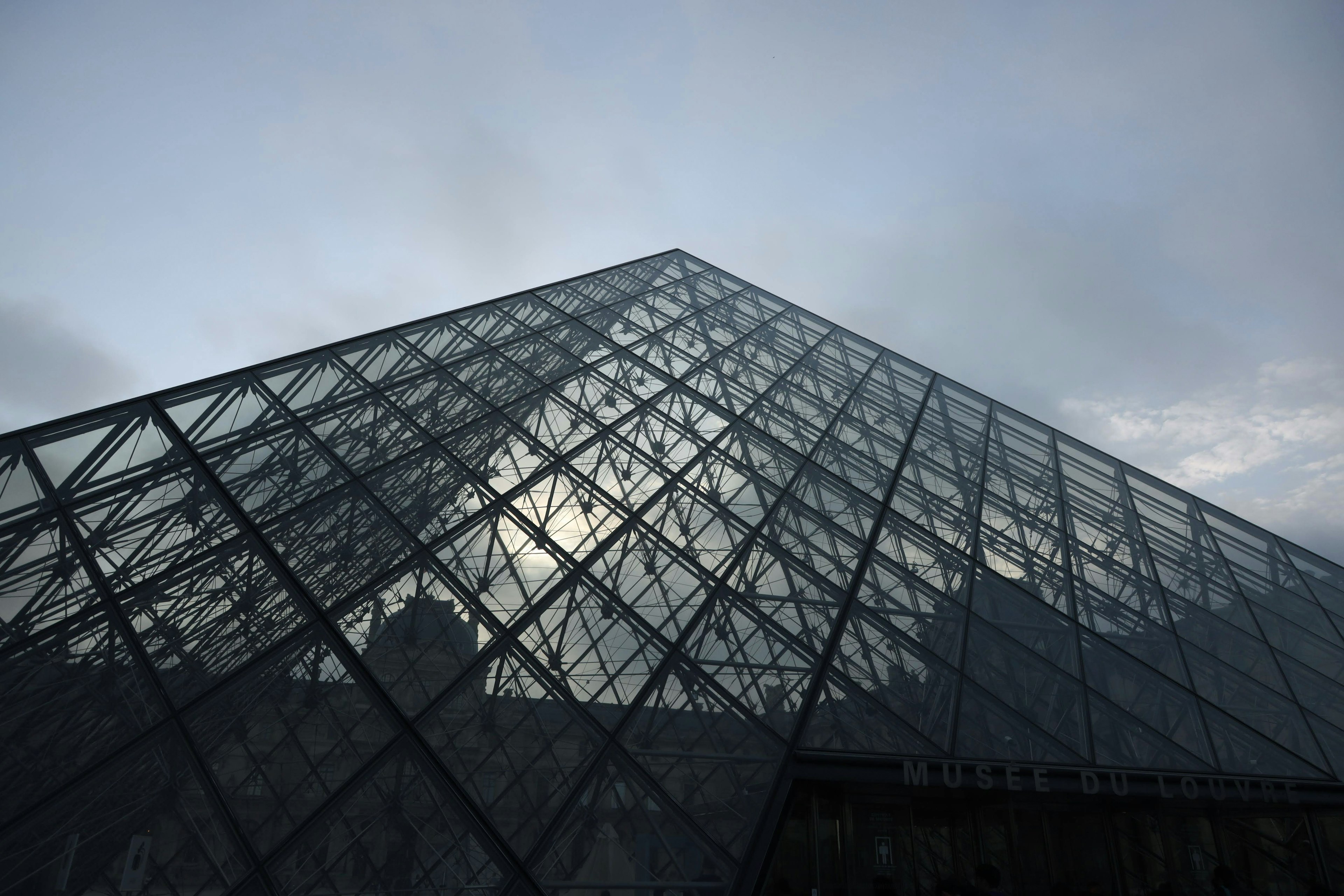 Vista diagonal de la pirámide de vidrio del Museo del Louvre con cielo nublado de fondo