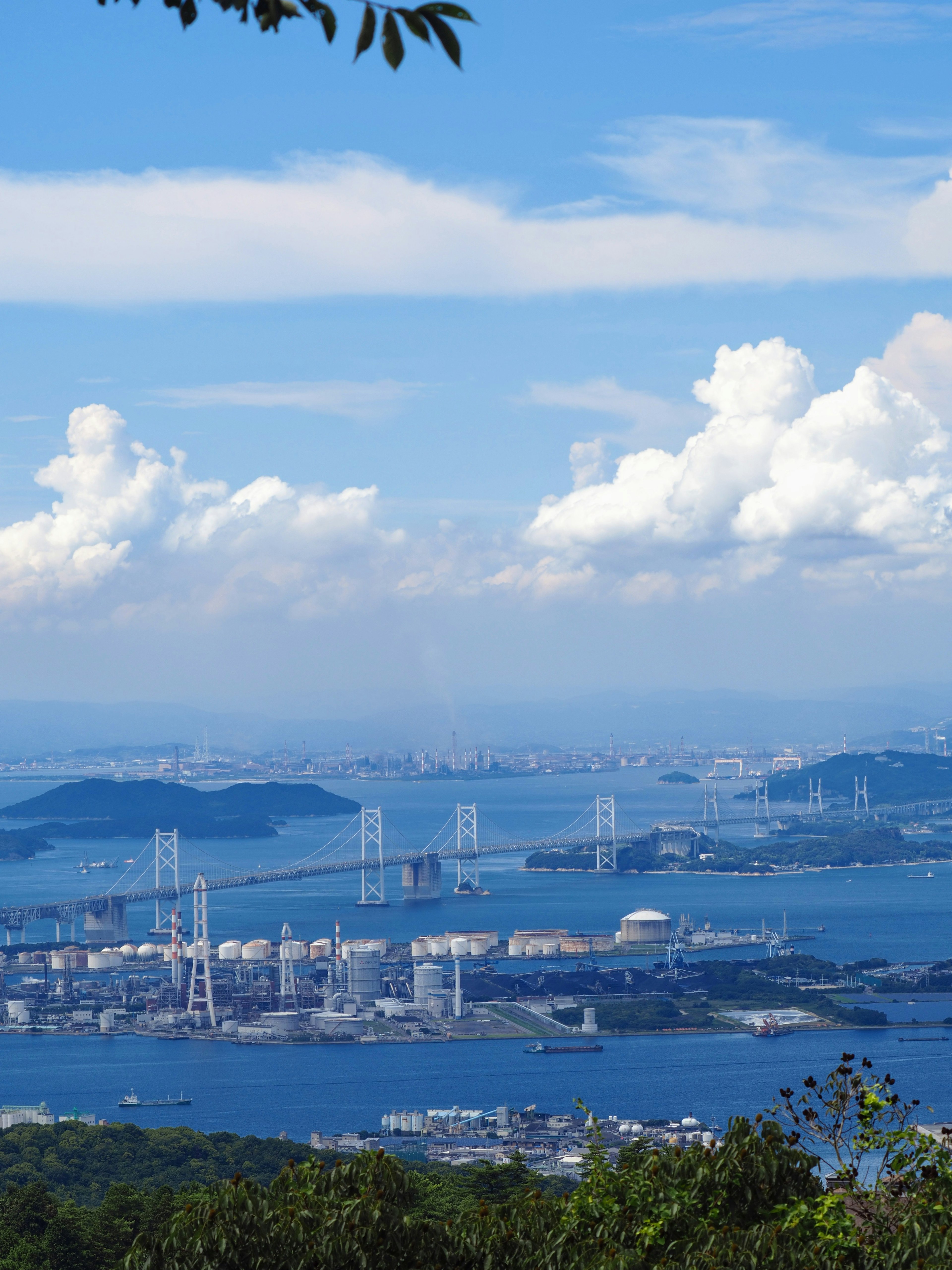 A panoramic view featuring blue sea and sky with white clouds industrial structures and bridges in the distance