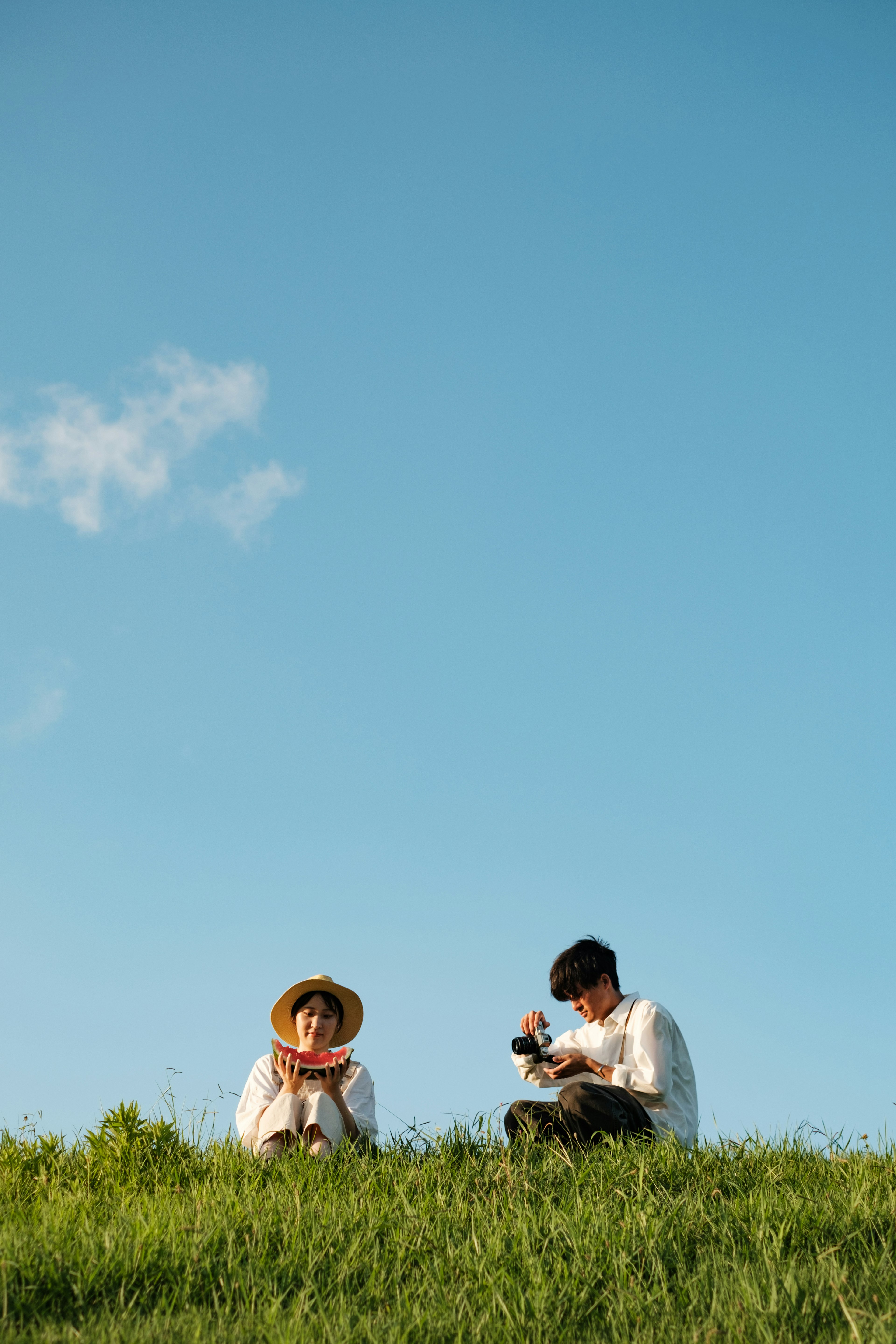 青空の下で草の上に座る二人の人物 一人はカメラを持ち もう一人はリラックスしている