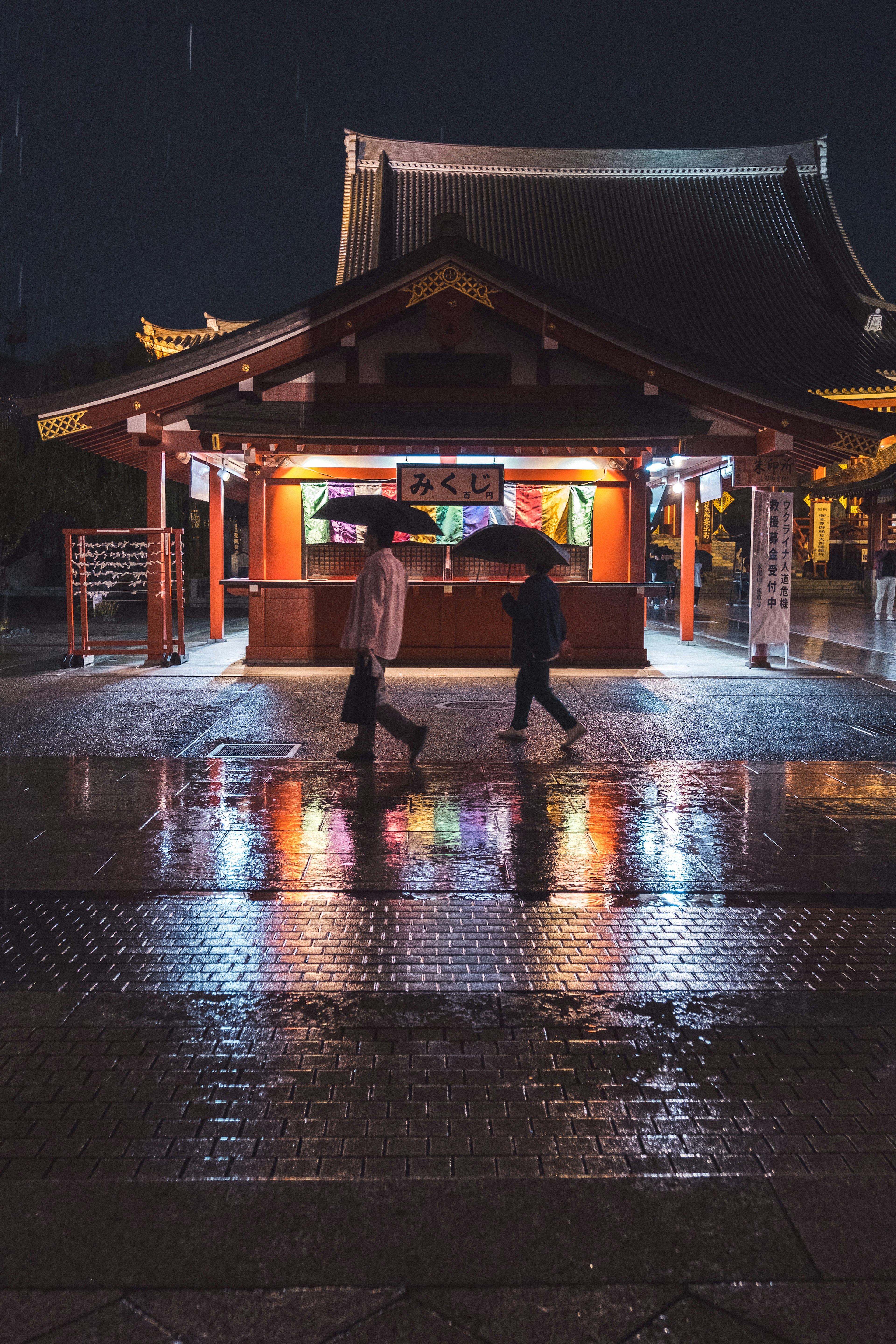 夜の雨の中で傘をさした二人が神社の前を歩いている