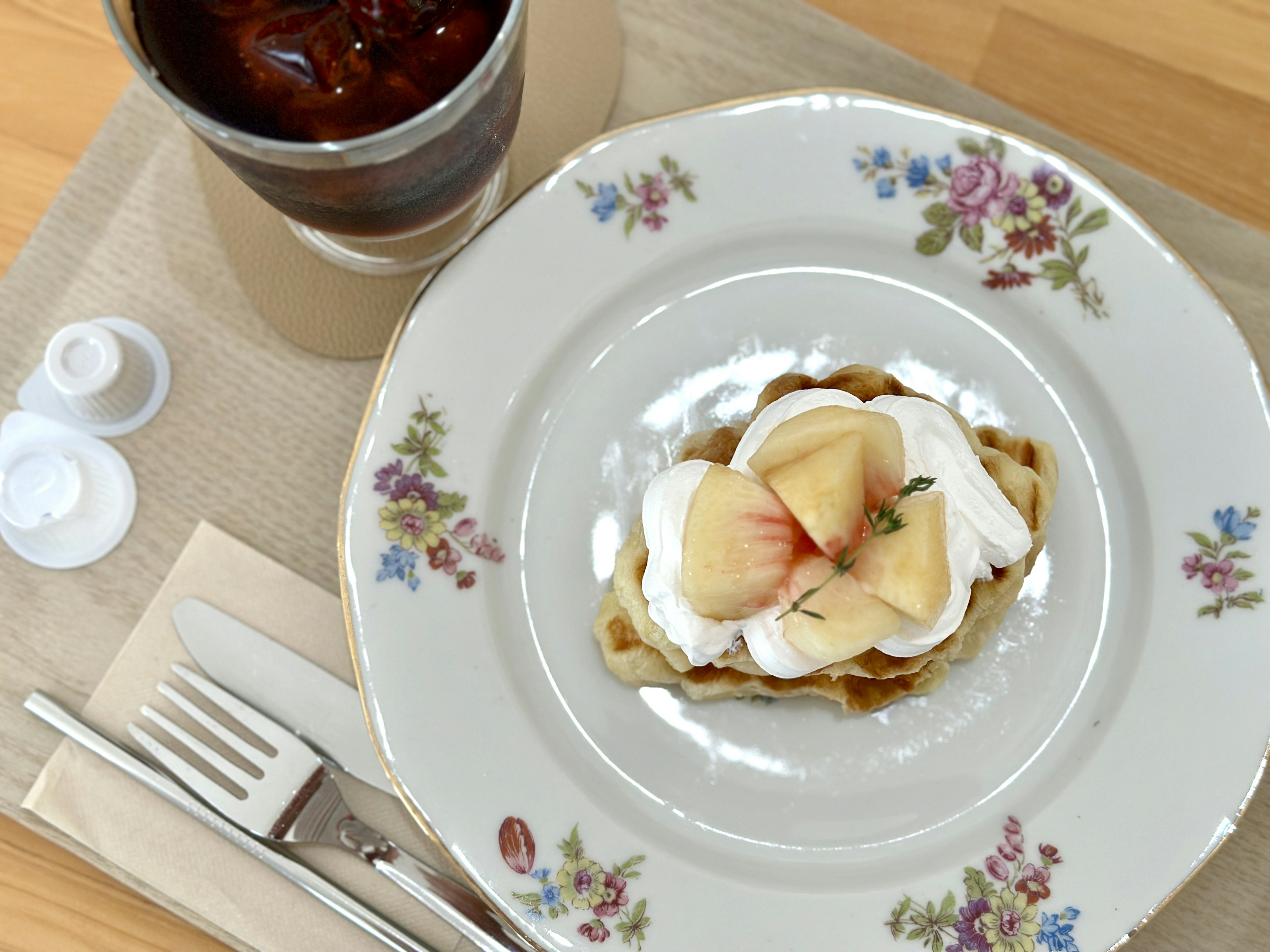 Un waffle cubierto con frutas y crema en un plato floral con café helado