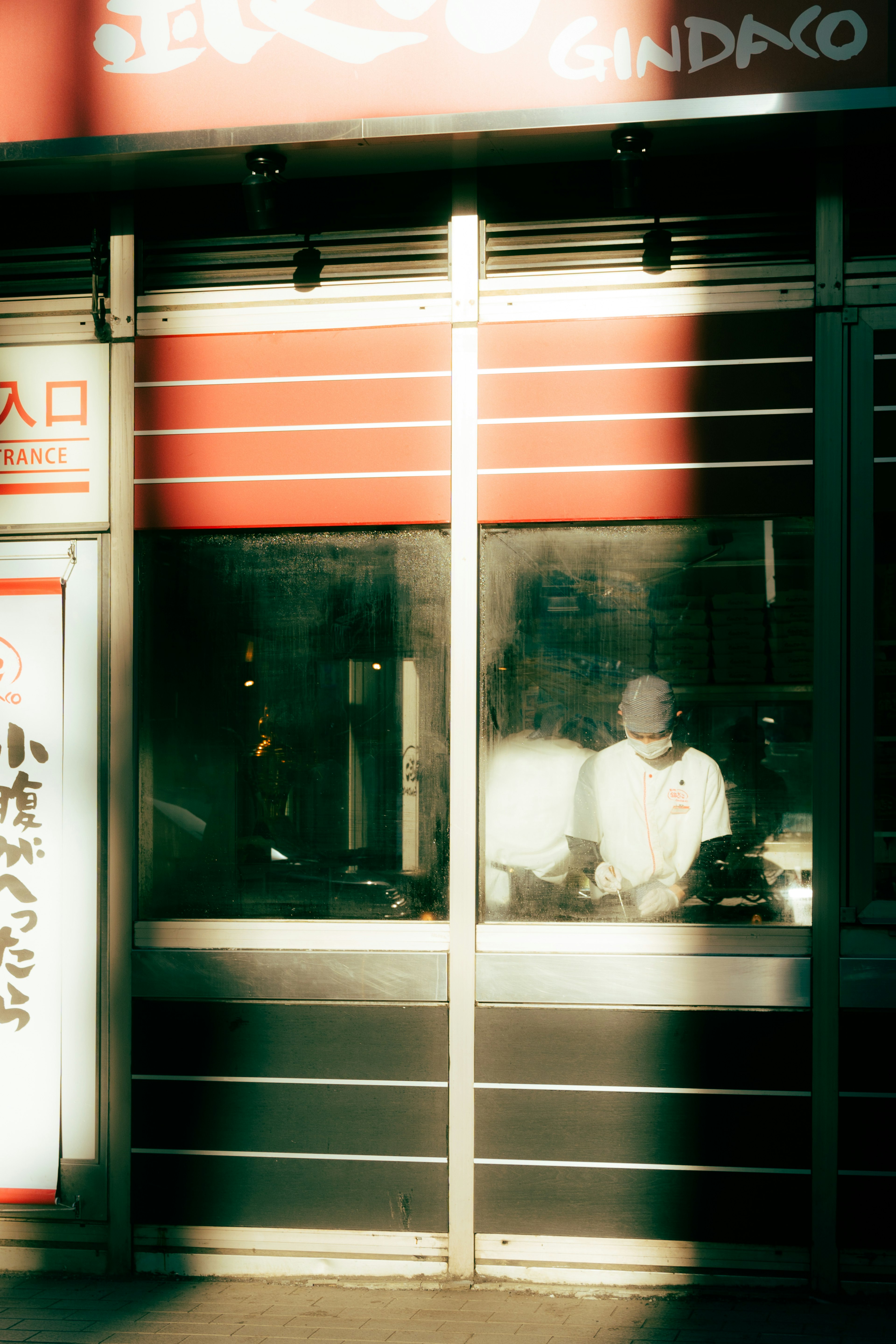 窓越しに料理を作るシェフと明るい赤い看板のある店舗