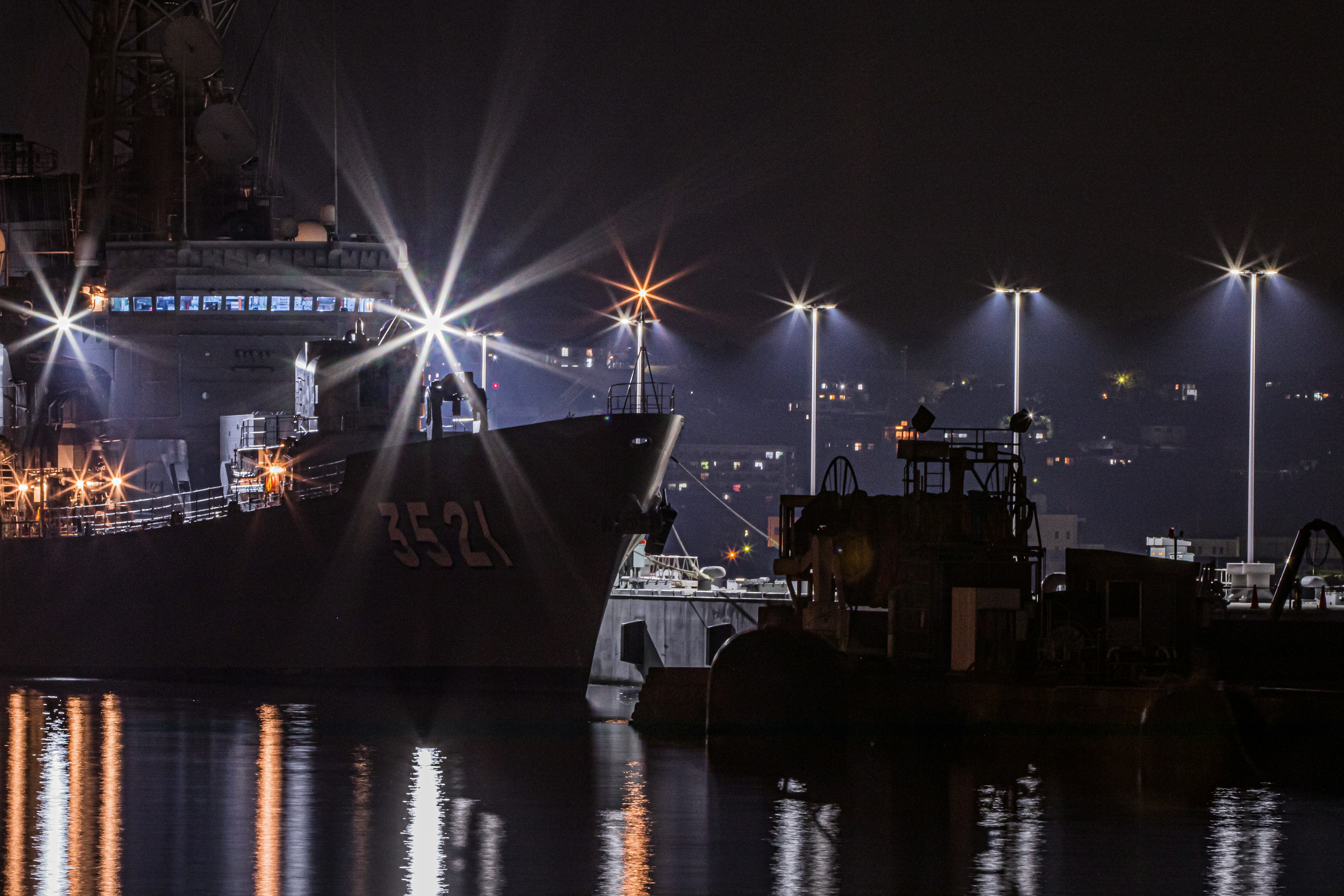 A military ship and a workboat docked at a harbor at night with bright lights