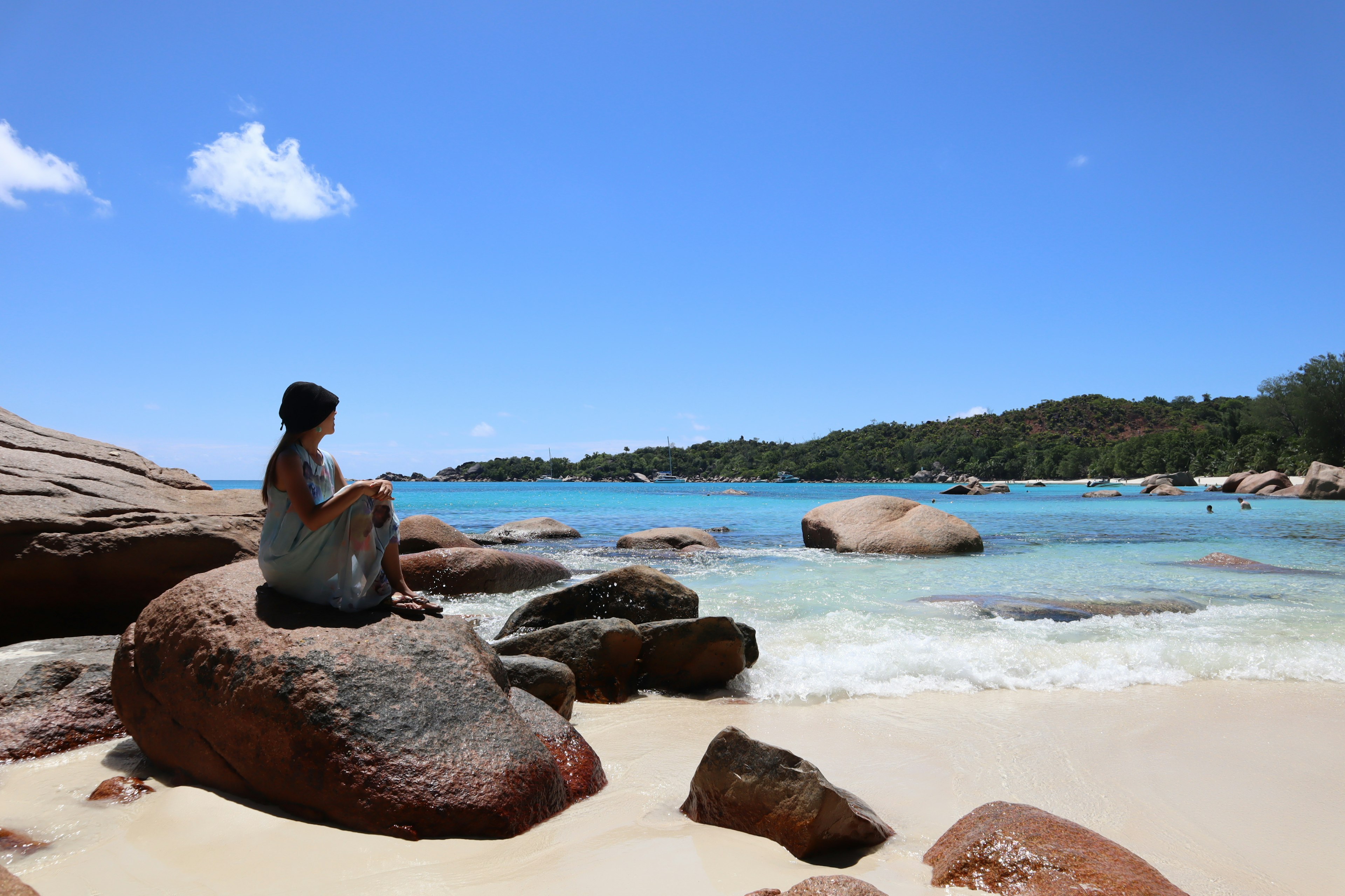 ผู้หญิงนั่งอยู่บนก้อนหินข้างทะเลสีน้ำเงินและชายหาดทรายขาว