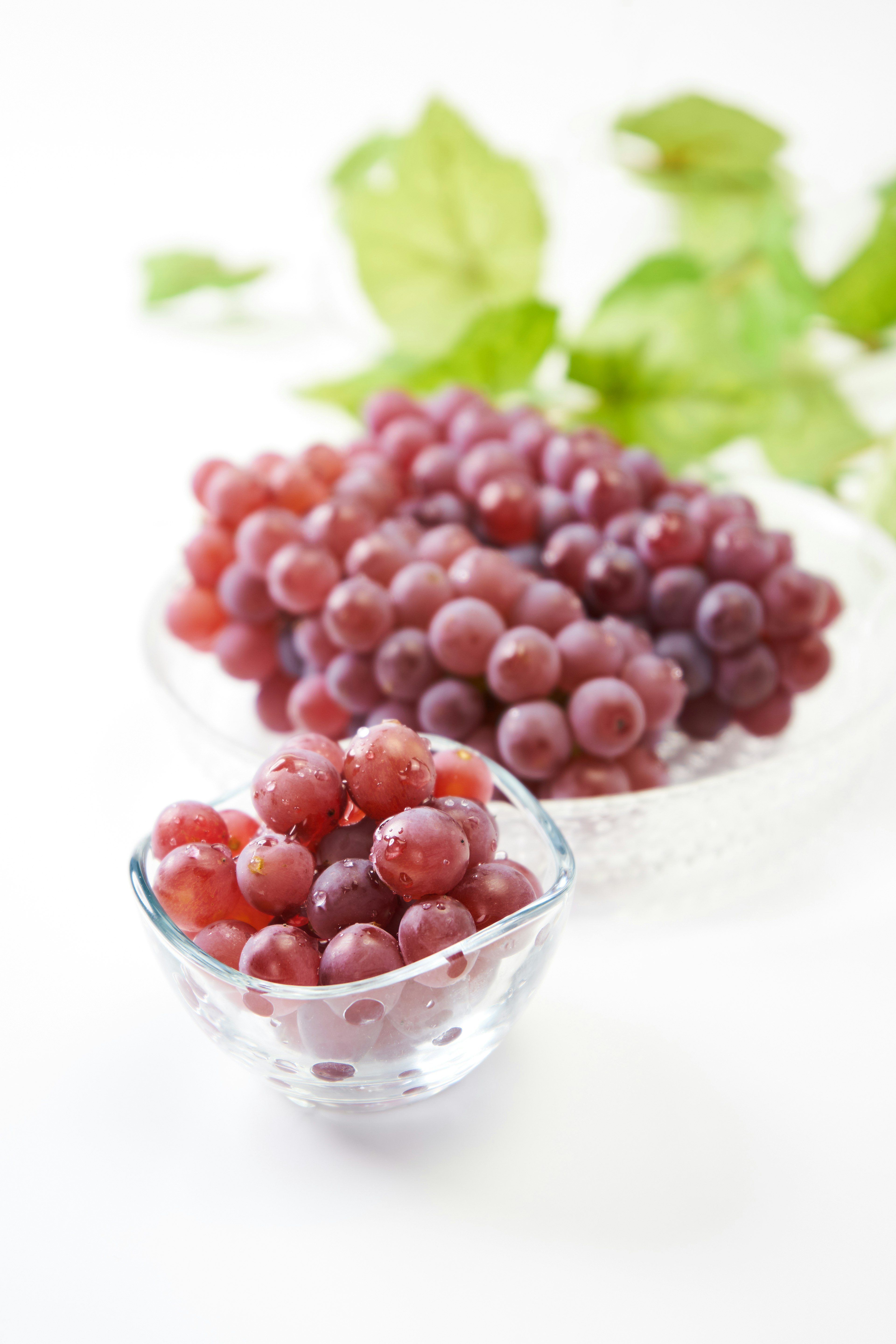 Imagen de uvas rojas en un tazón de vidrio y un tazón más pequeño con hojas verdes de fondo