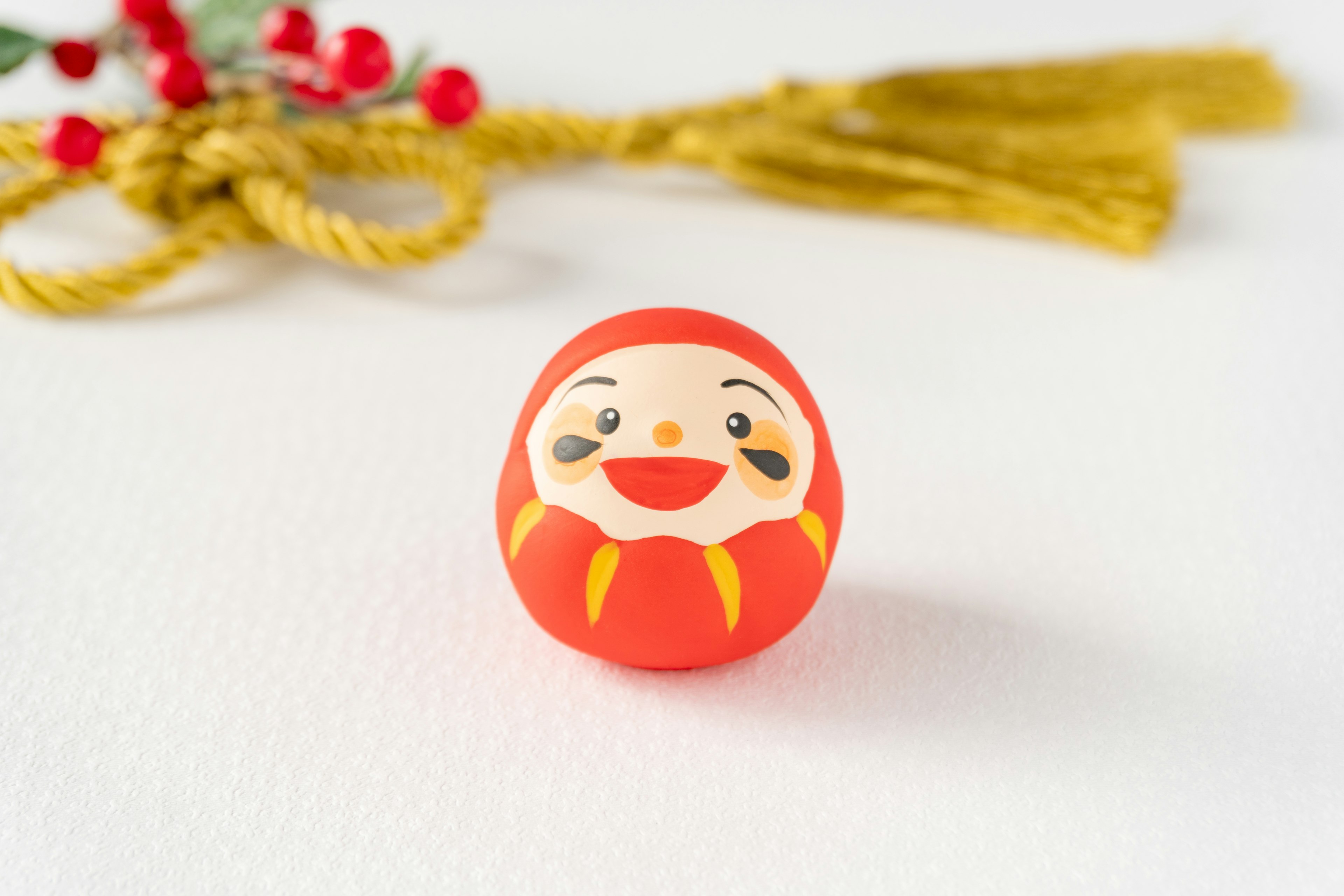 A red Daruma doll in the center with golden tassels and red berries in the background