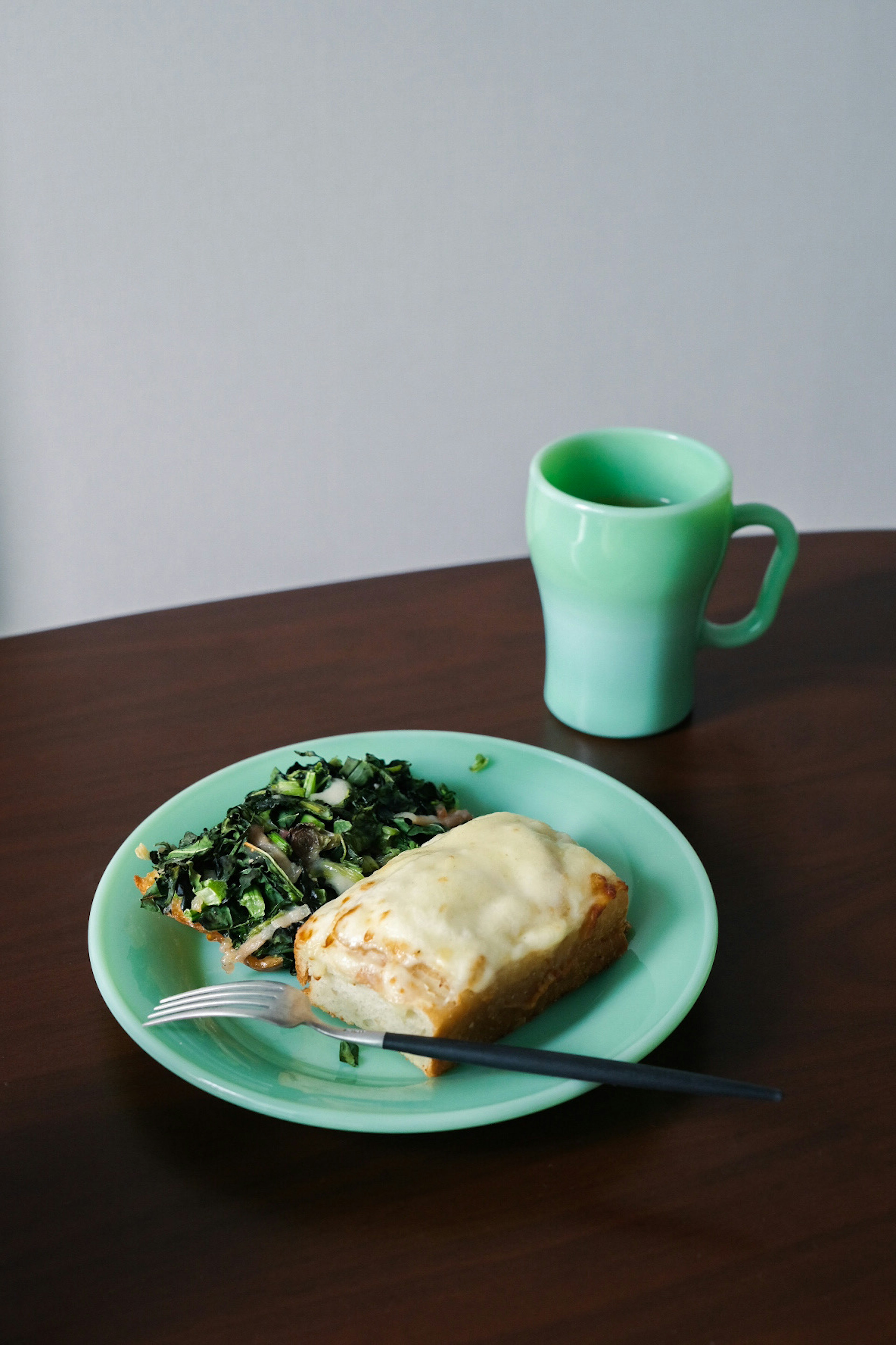 A meal on a green plate featuring a cheese-topped pastry and spinach salad with a green mug on a wooden table