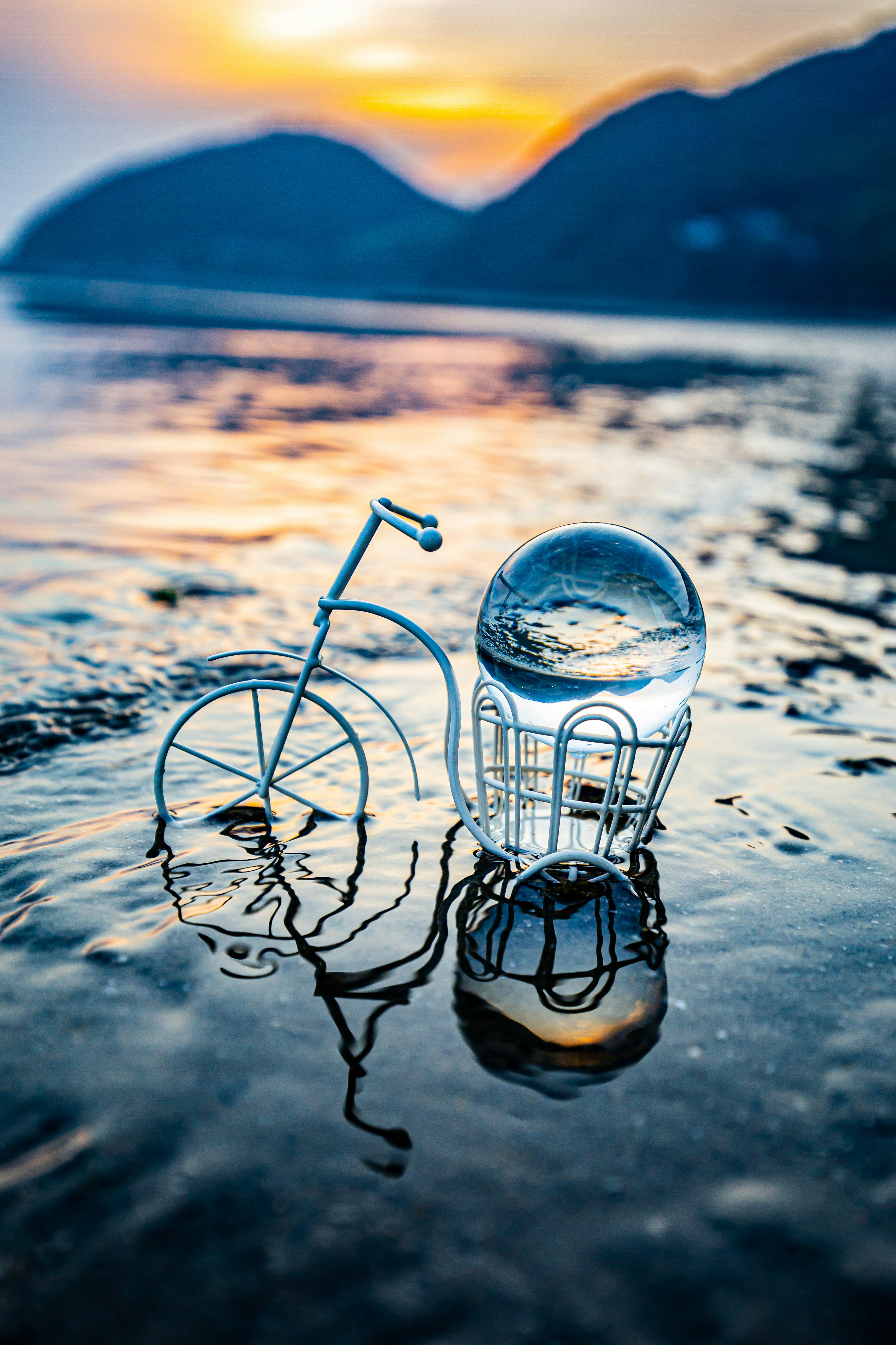 A bicycle model and a glass orb reflecting on water at sunset with mountains in the background