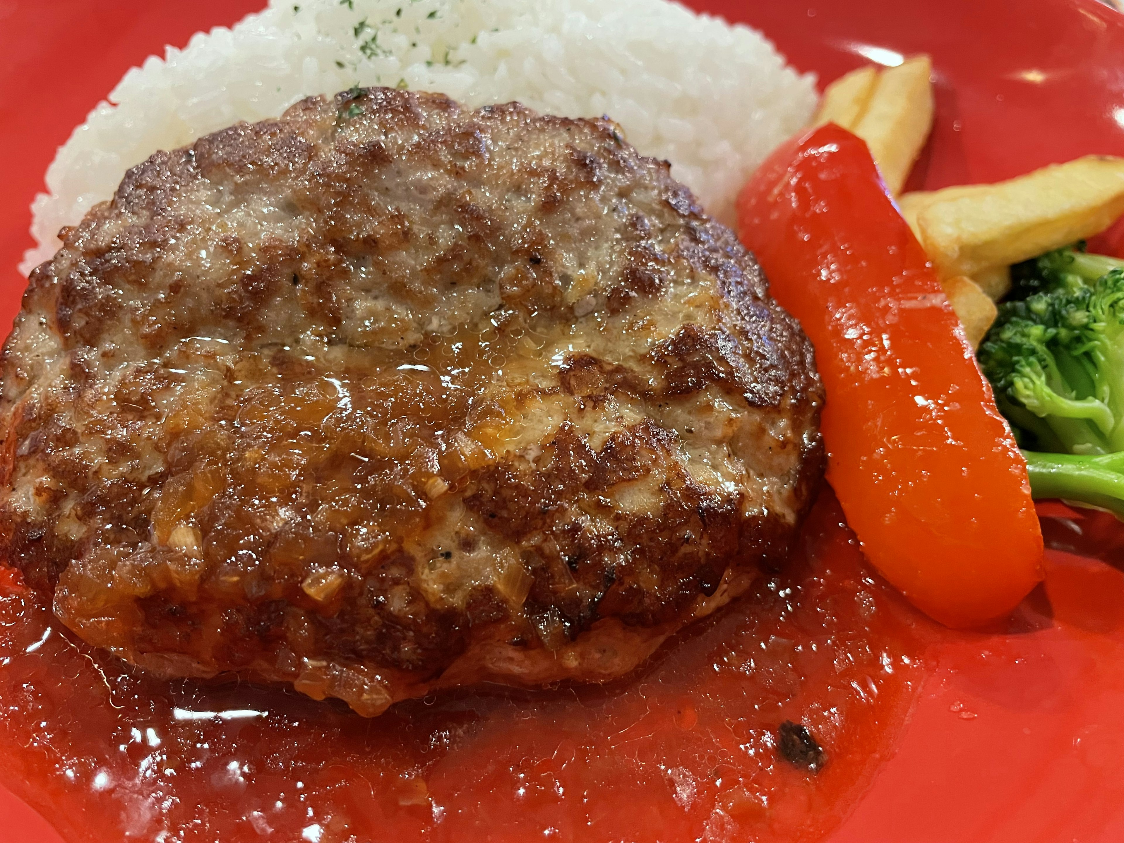 Un steak haché servi sur une assiette rouge avec du riz des brocolis et un poivron rouge