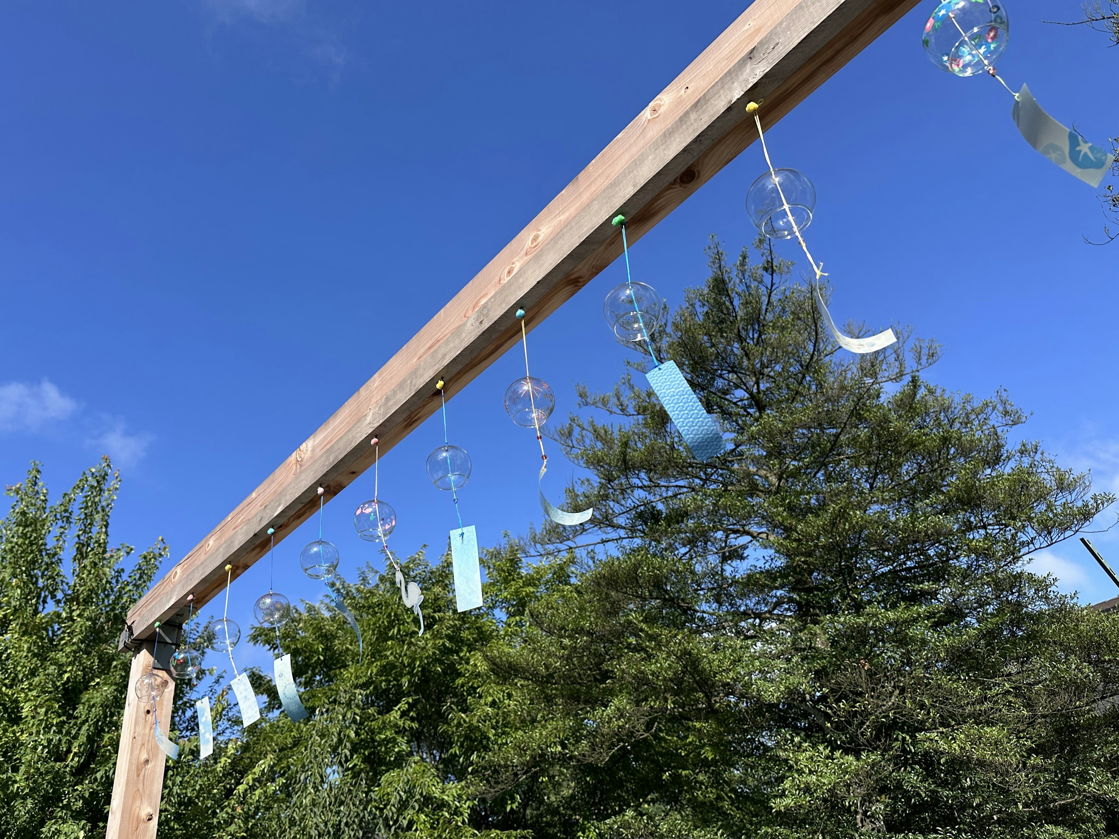 Wind chimes art installation hanging under blue sky wooden frame with green trees in the background