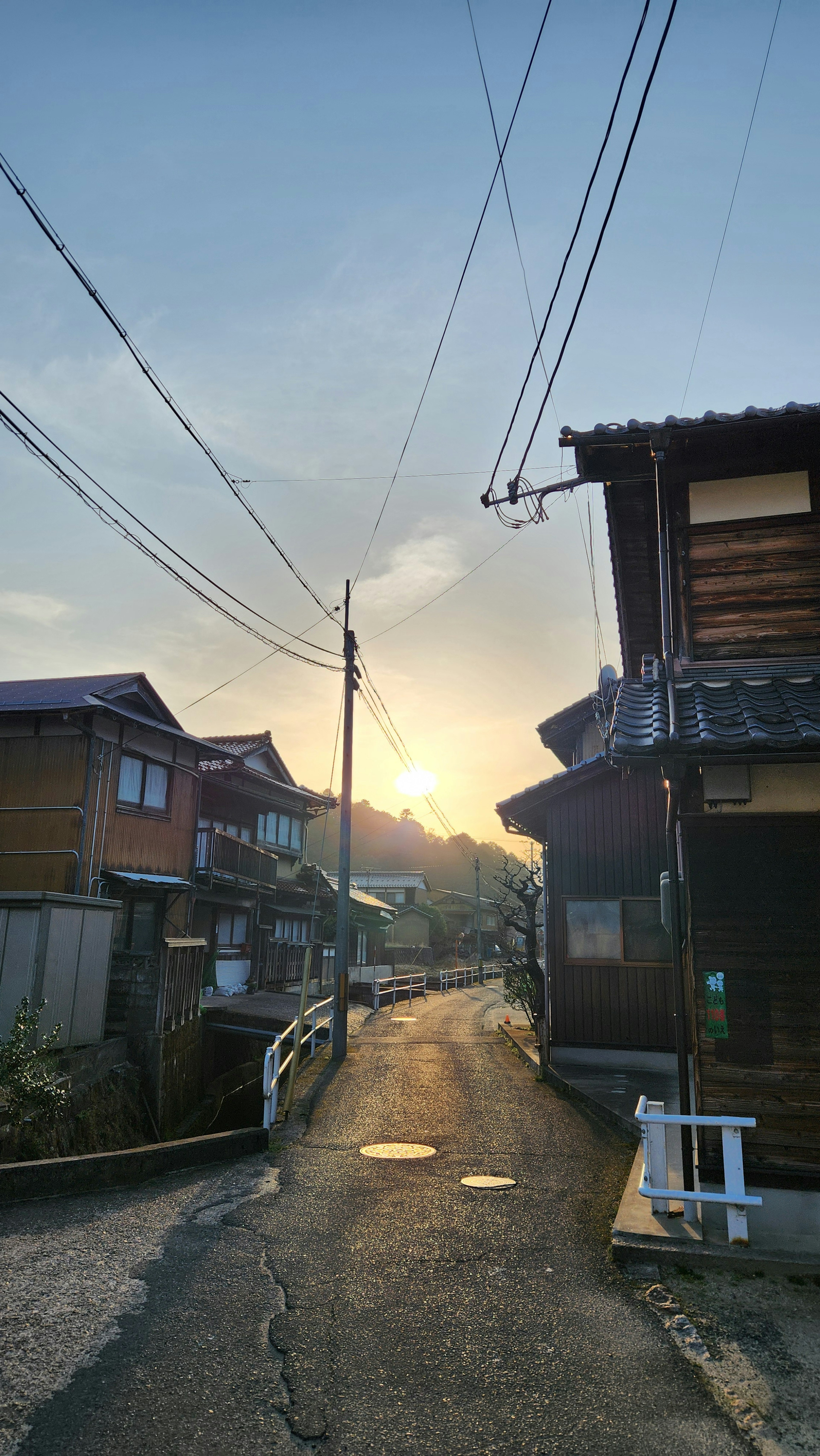 静かな住宅街の夕暮れ時の風景で、木造の家々が並び、空には夕日が映える