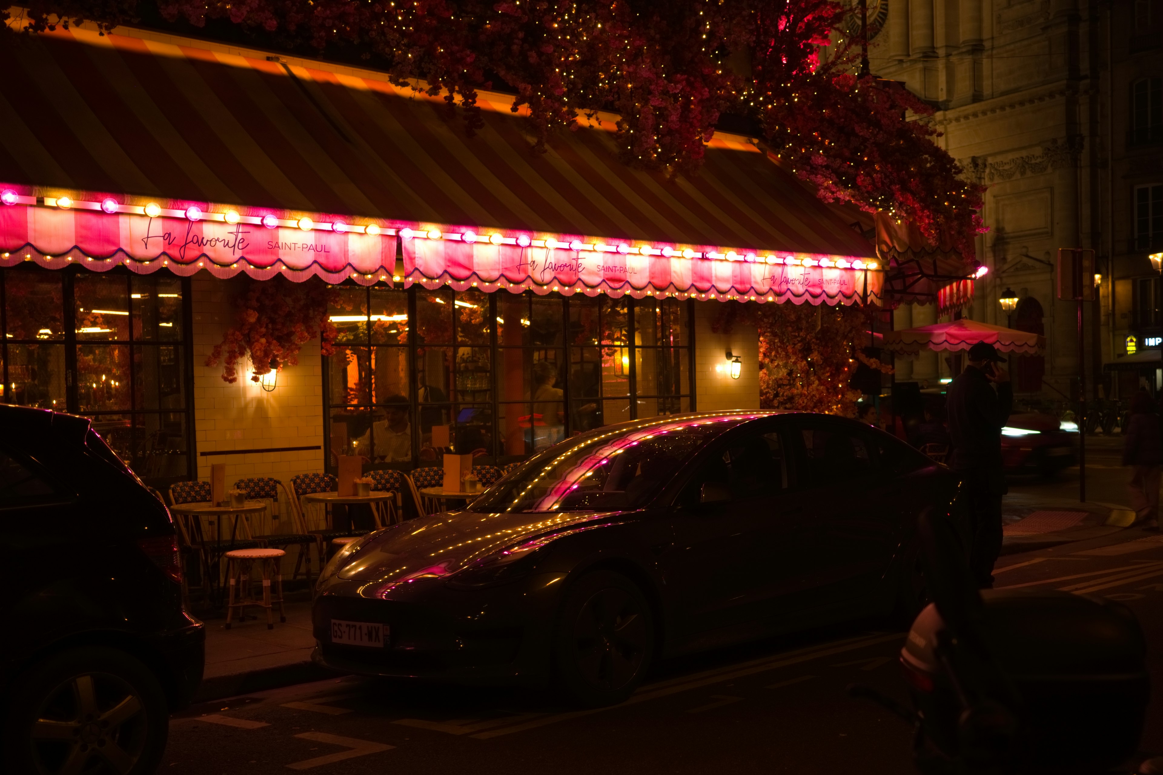 Exterior de un café iluminado por luces rosas en una escena nocturna con un coche negro