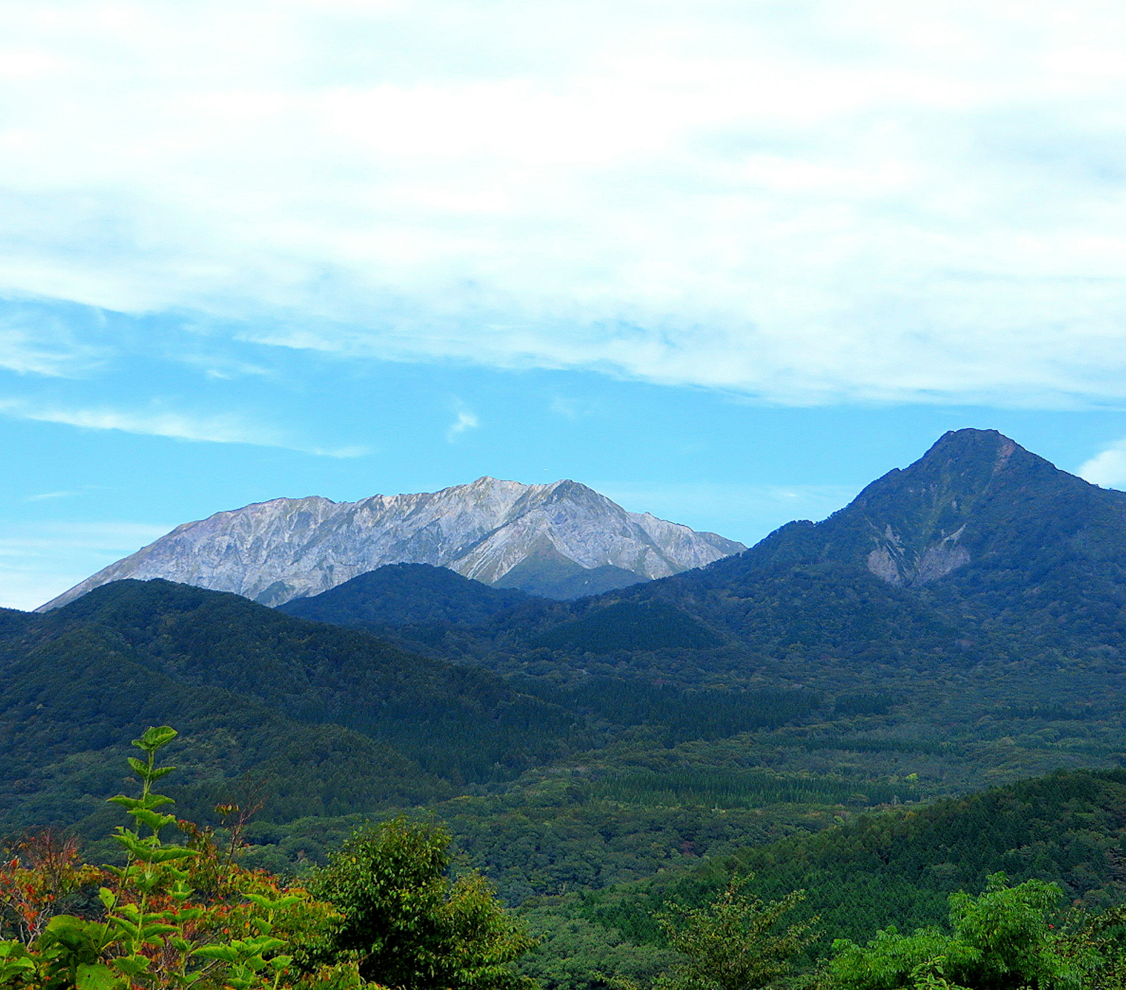 ภูเขาสีเขียวใต้ท้องฟ้าสีฟ้าพร้อมยอดเขาสีเทา