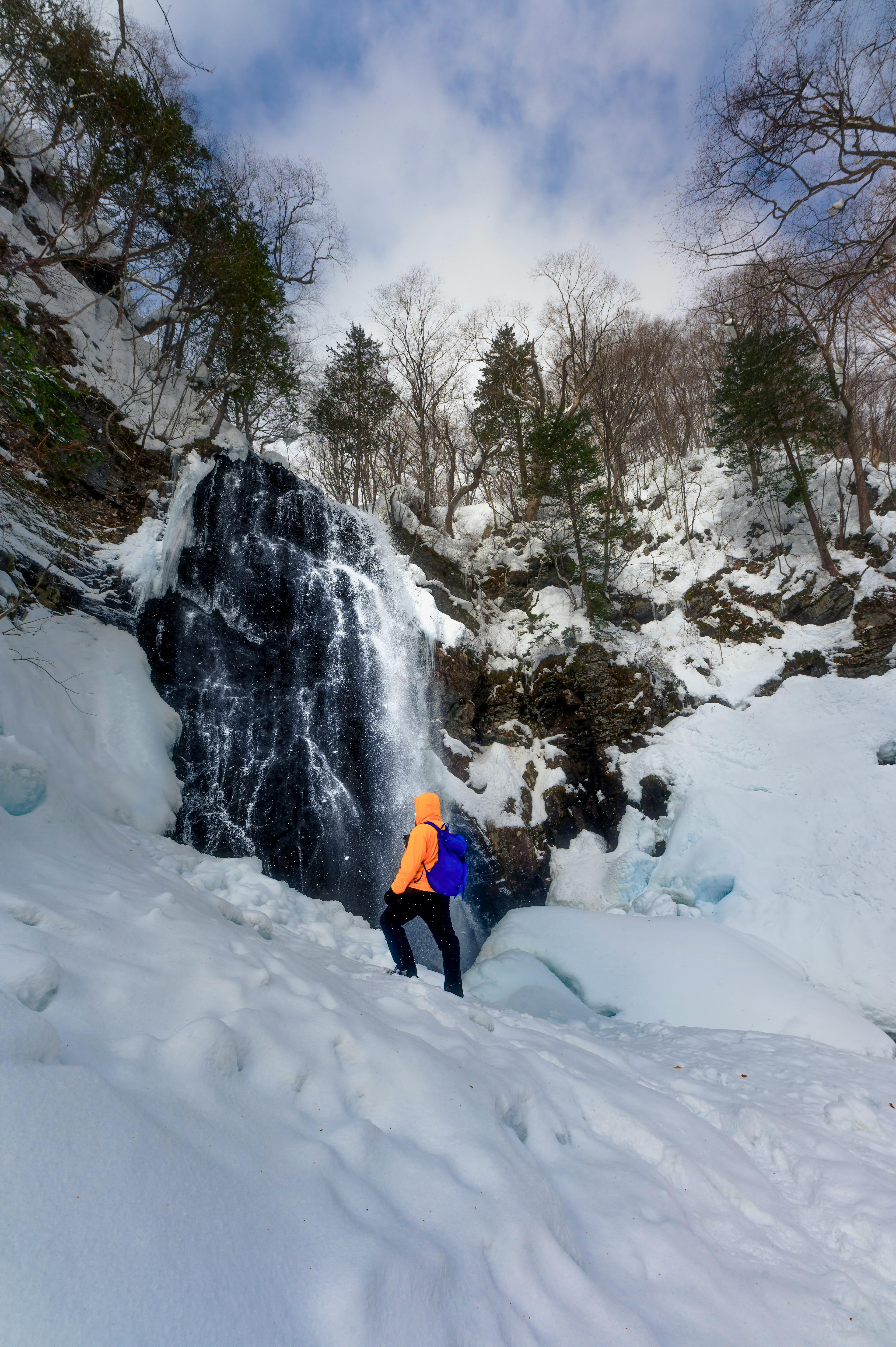 Wanderer in orange Jacke vor einem gefrorenen Wasserfall in schneebedeckter Landschaft