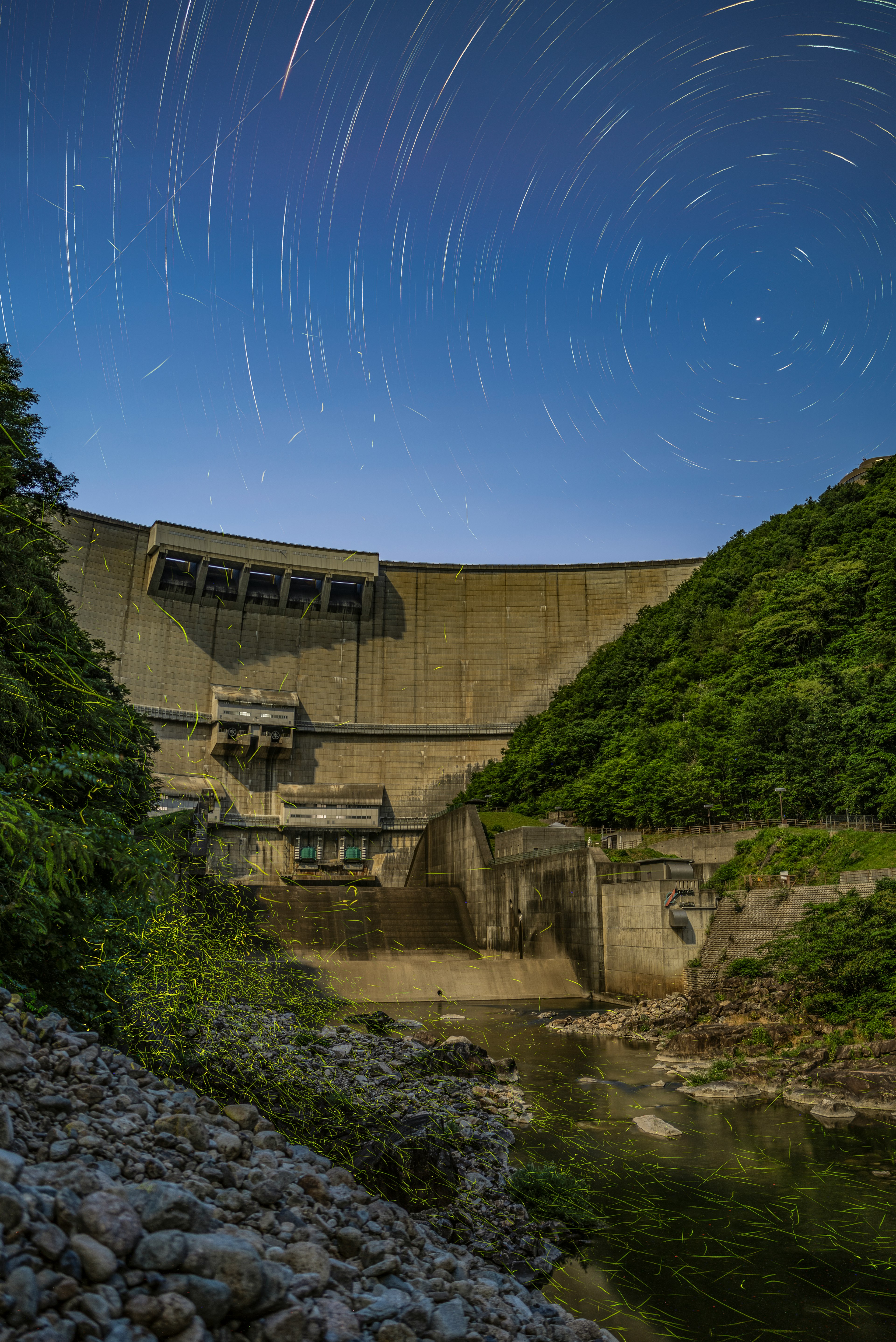 星の軌跡が描かれた夜空の下にあるダムの風景 緑豊かな丘に囲まれたコンクリート構造物