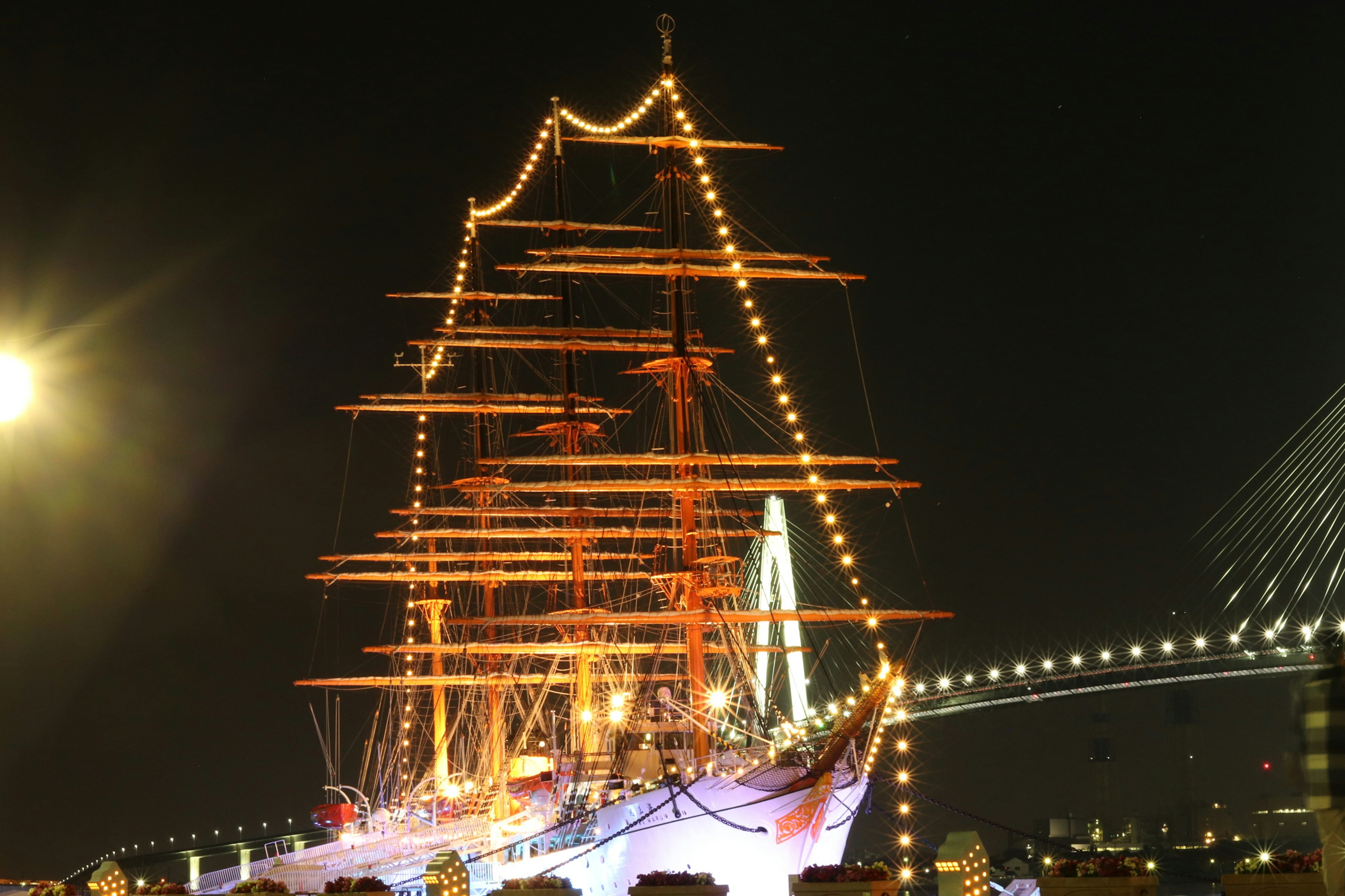 Illuminated tall ship against a night sky with a bridge silhouette