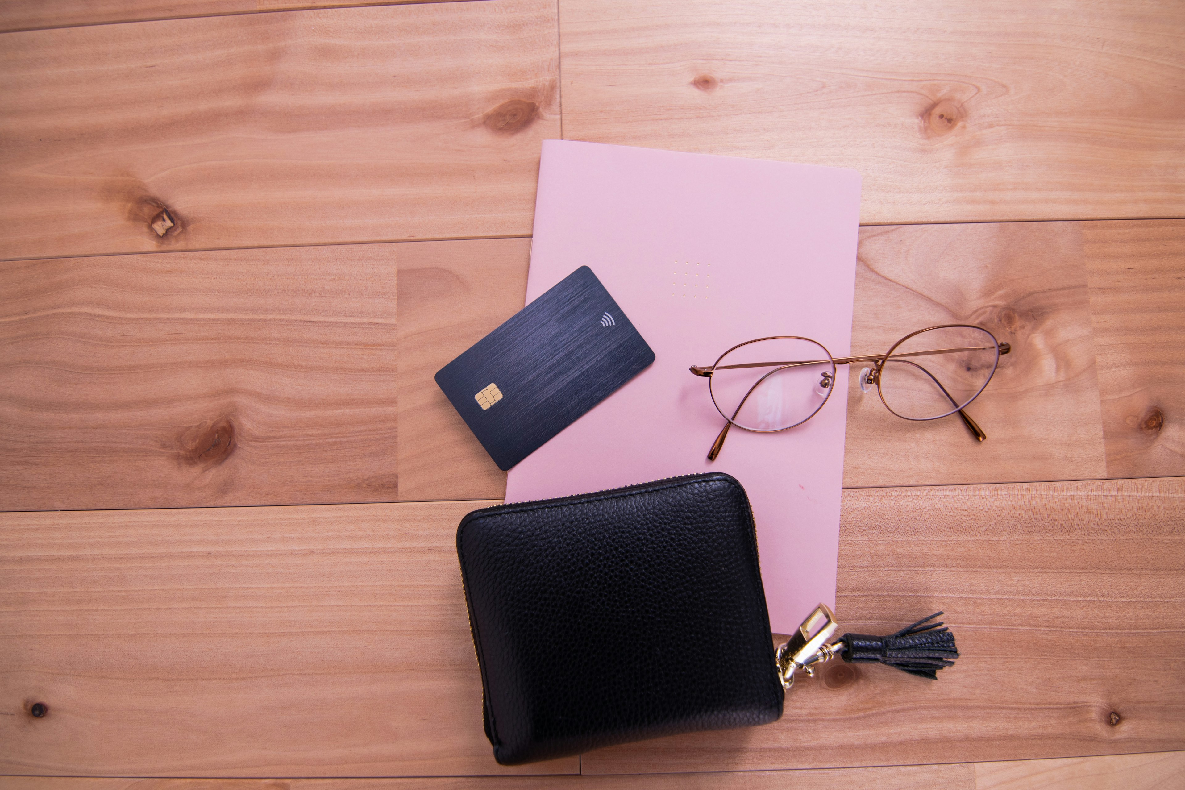 A black wallet, pink notebook, credit card, and glasses on a wooden table