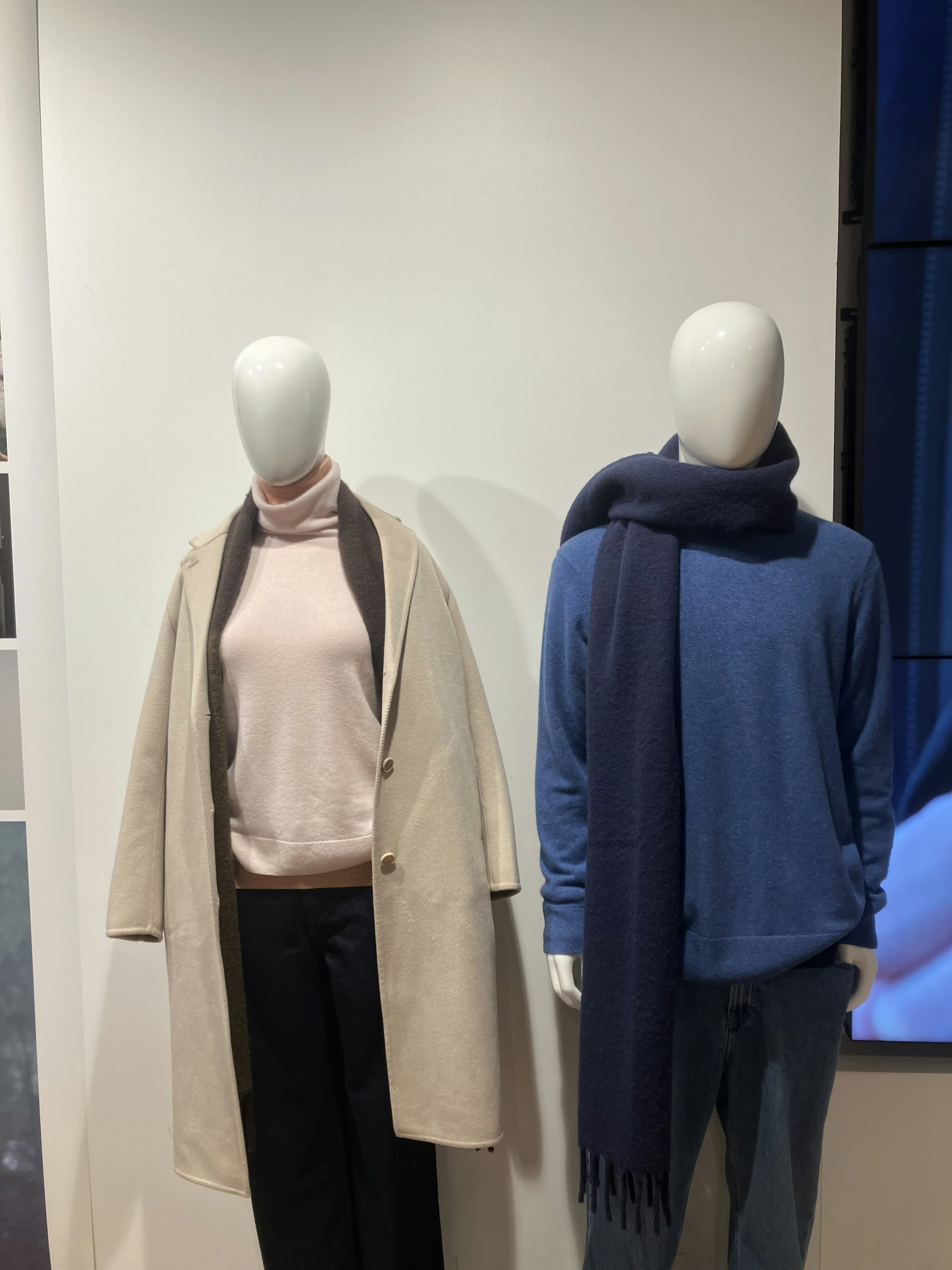 Display of mannequins in a fashion store featuring a cream coat and pink sweater on the left mannequin and a blue sweater with navy scarf on the right mannequin