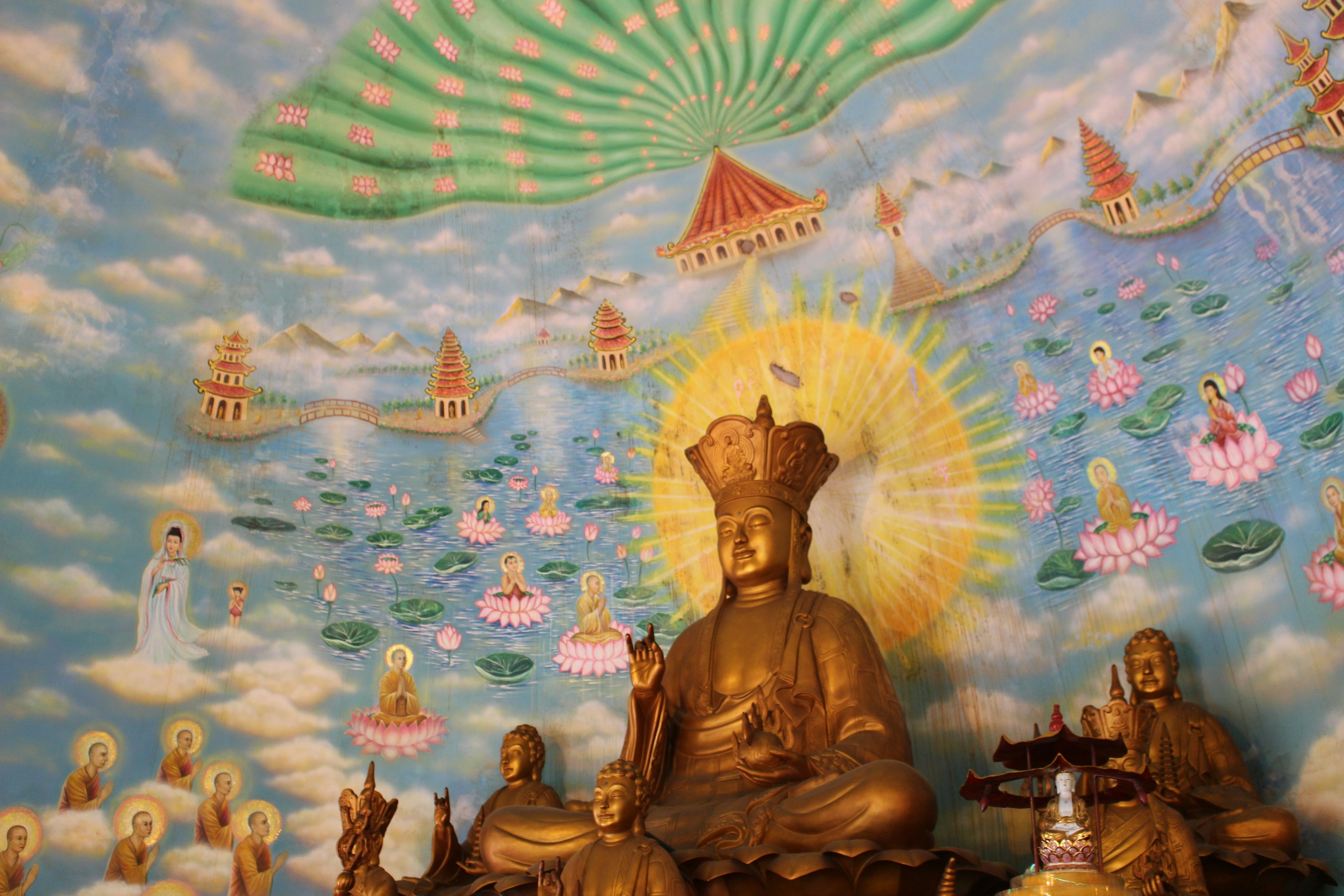 Buddha statue in front of a vibrant Buddhist mural featuring clouds and lotus flowers