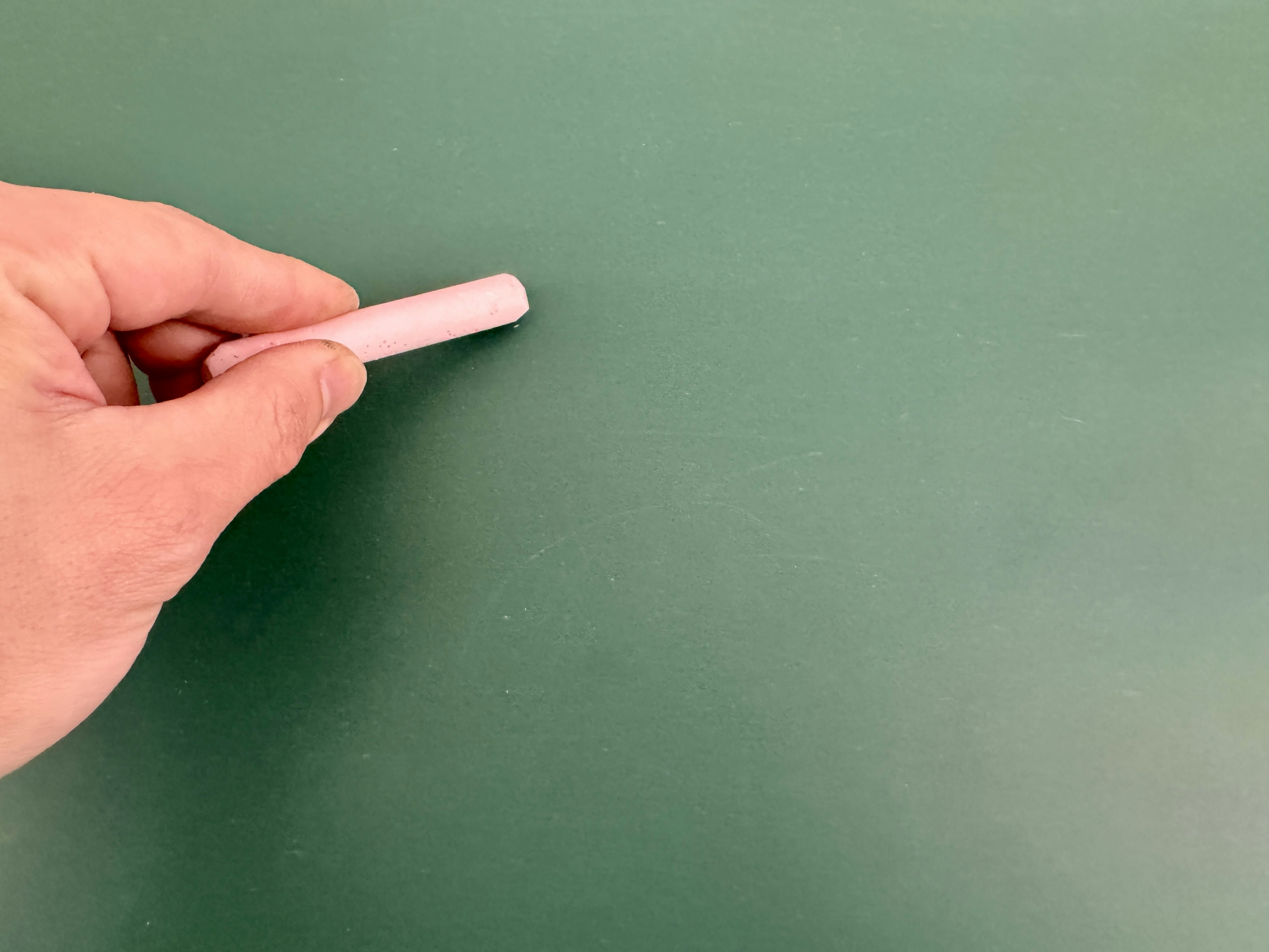 A hand holding a white chalk touching a green chalkboard