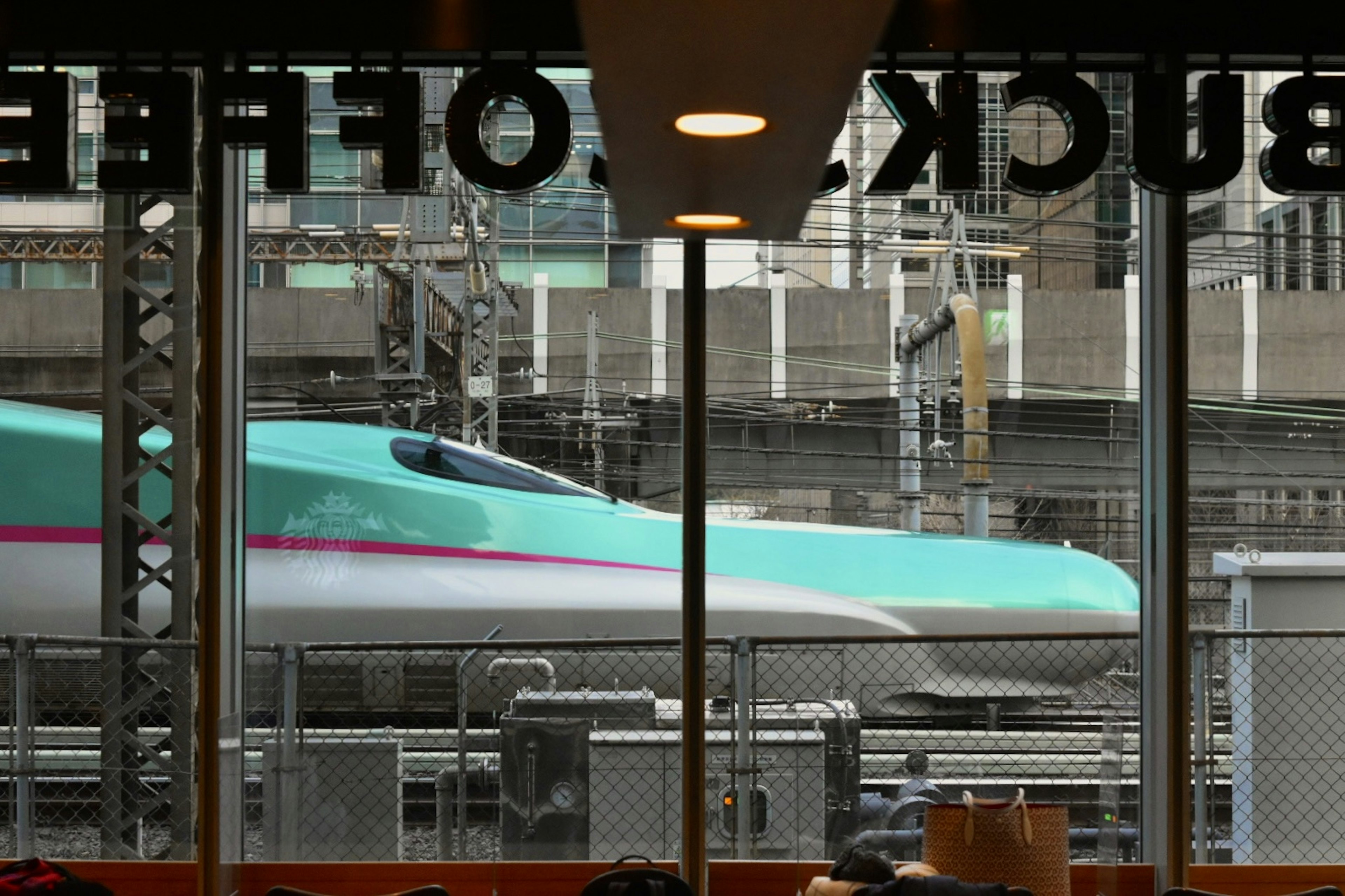 Pemandangan kereta Shinkansen dengan desain hijau dan pink dari jendela kafe