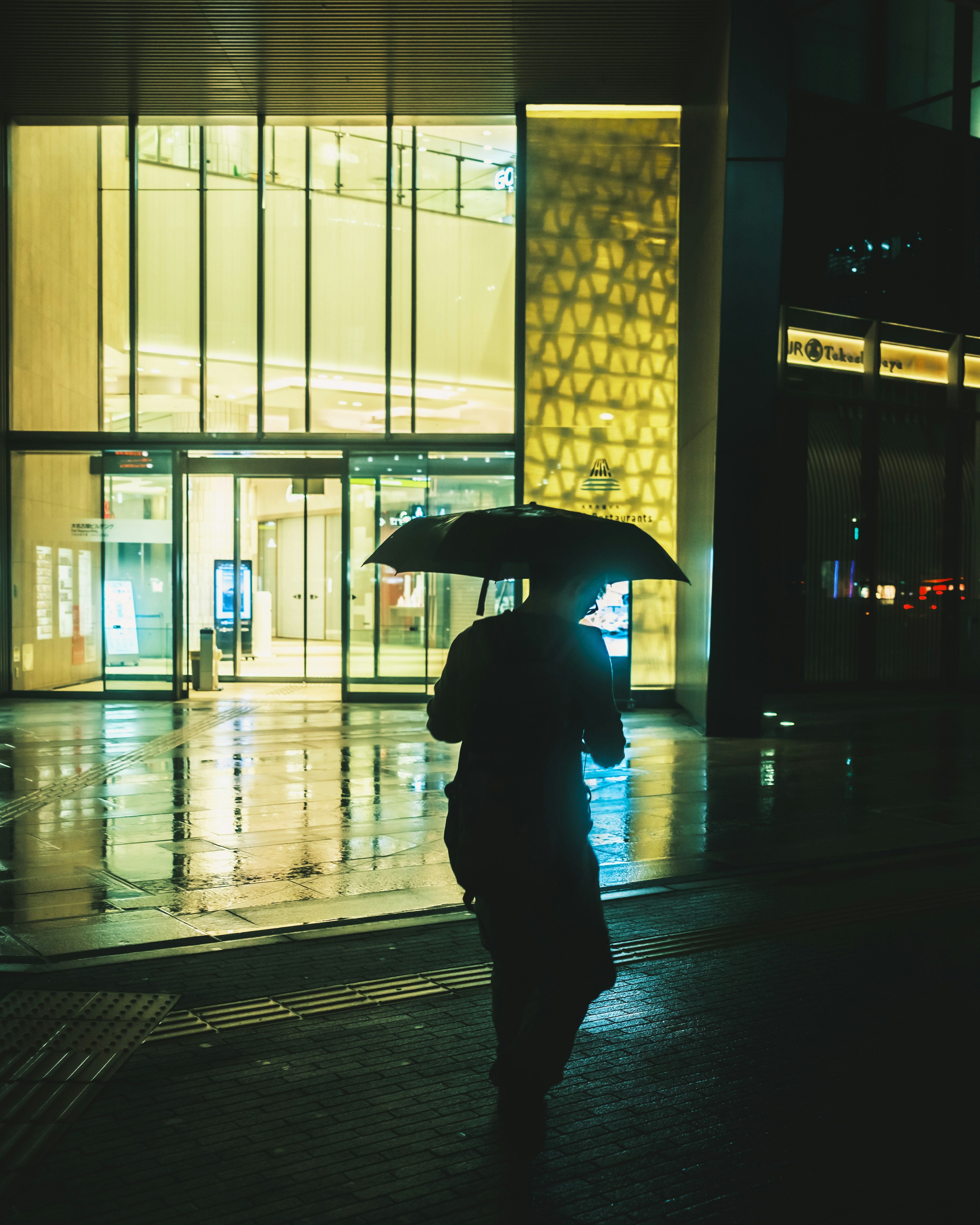 雨の中で傘を持った人物が現代的な建物の前を歩いている