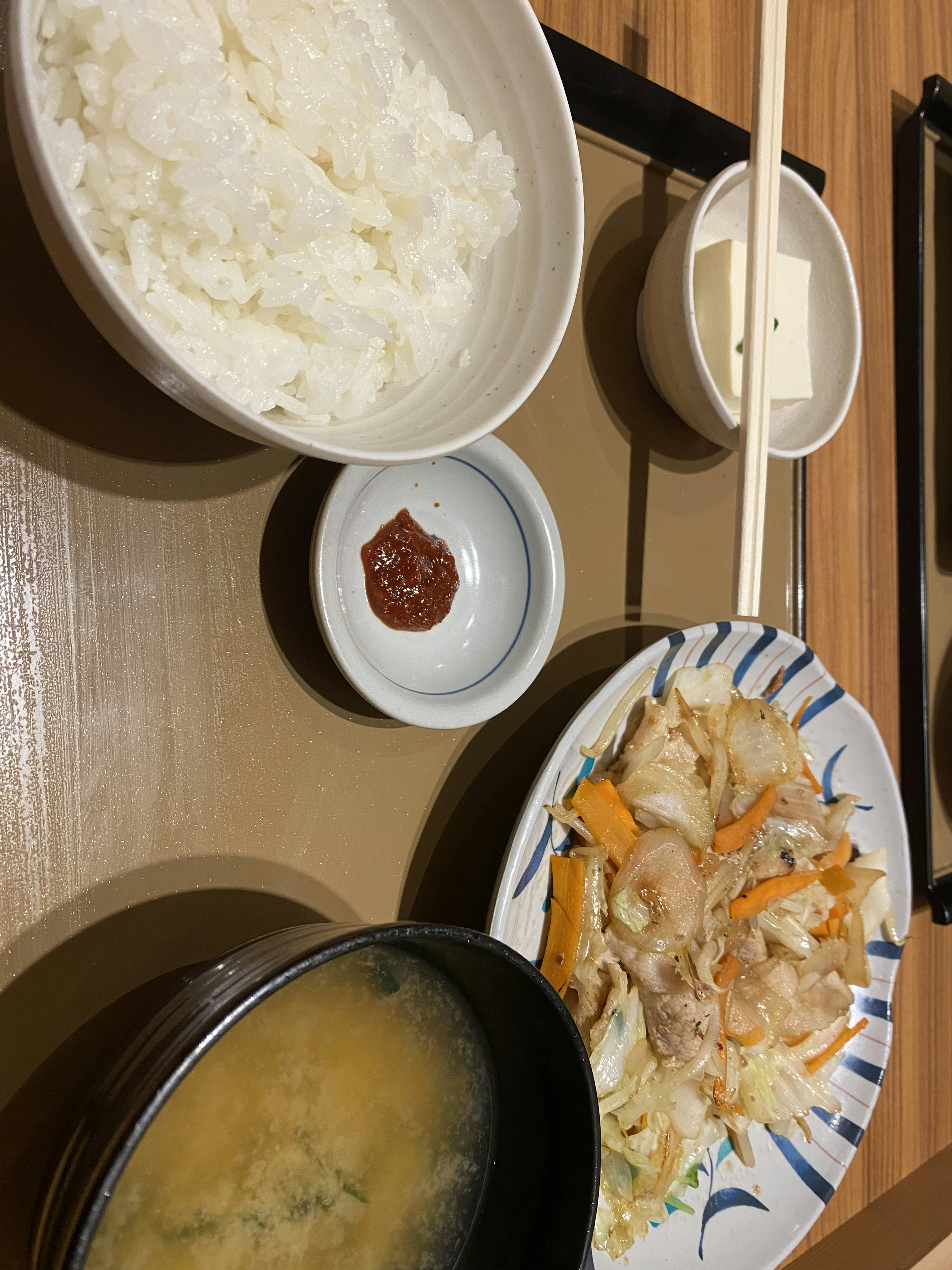 A Japanese meal featuring rice, miso soup, a dish of vegetables and pork, tofu, and a small bowl of condiment