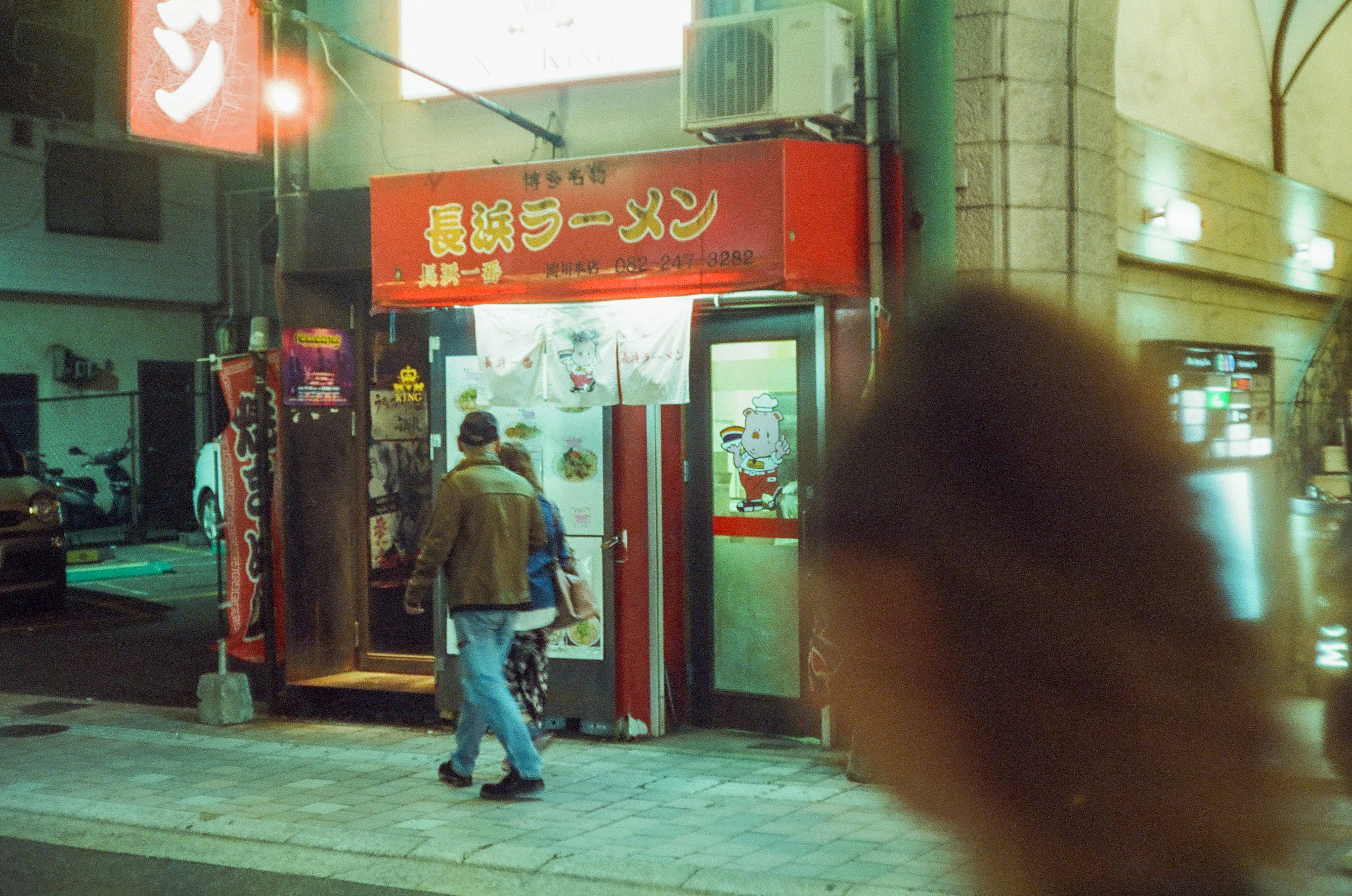 Night scene of a ramen shop with a red sign and people walking by