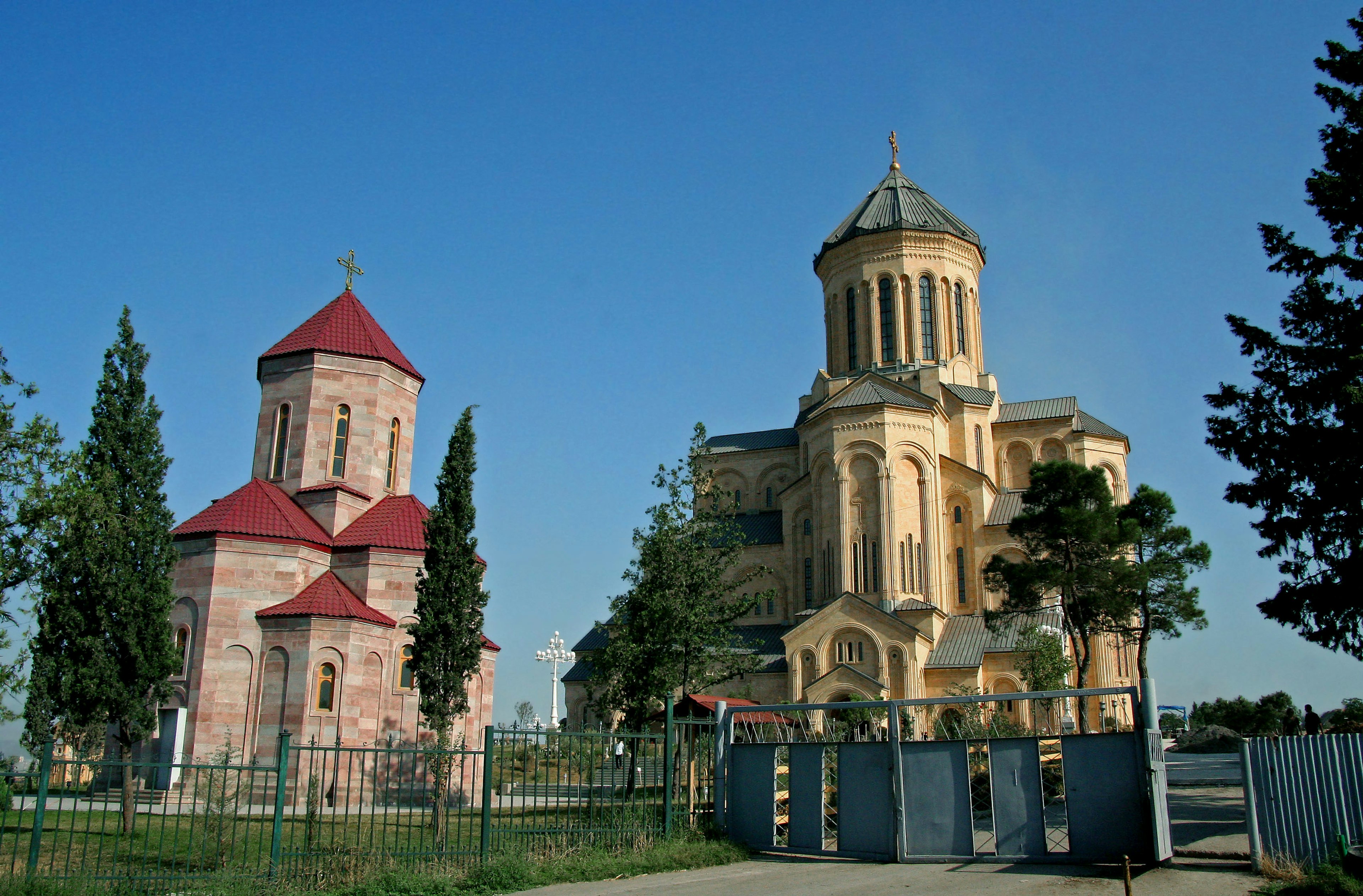 Two church buildings under a blue sky surrounded by green trees featuring a small church with a red roof and a large golden church