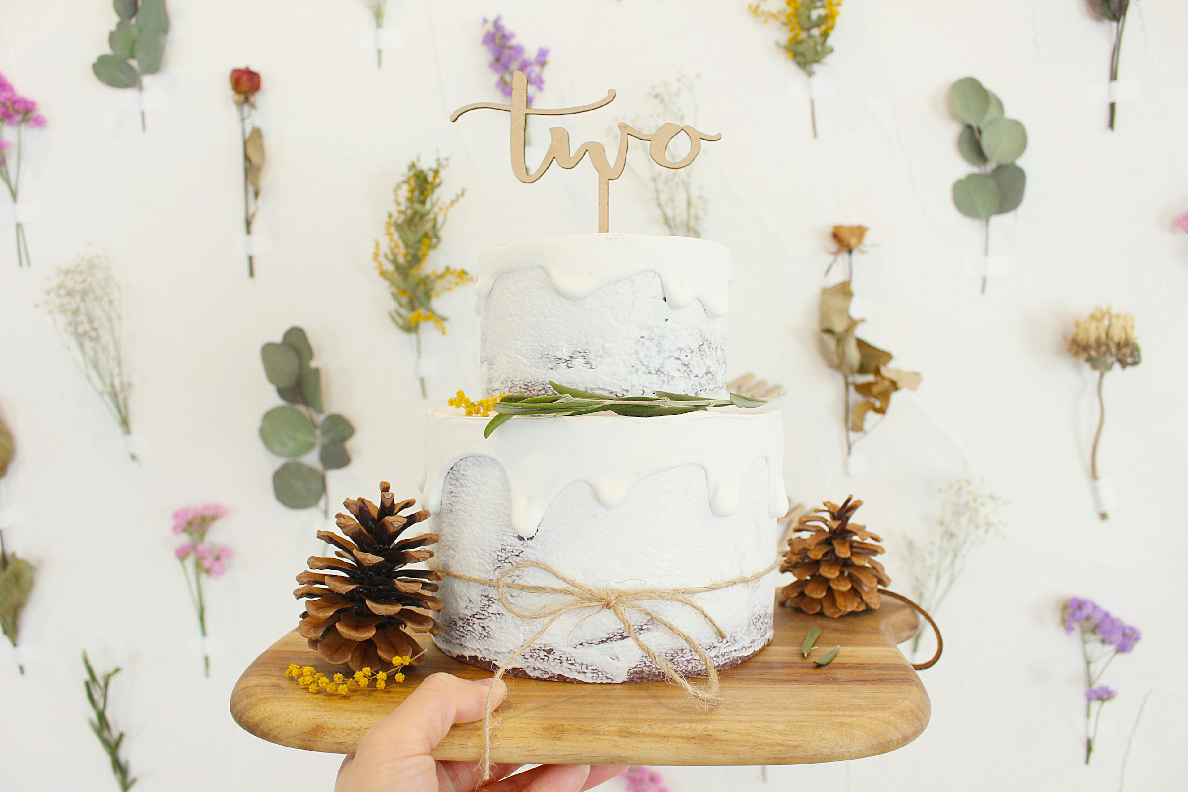 Gâteau blanc à deux étages sur un plateau en bois avec des fleurs séchées en arrière-plan