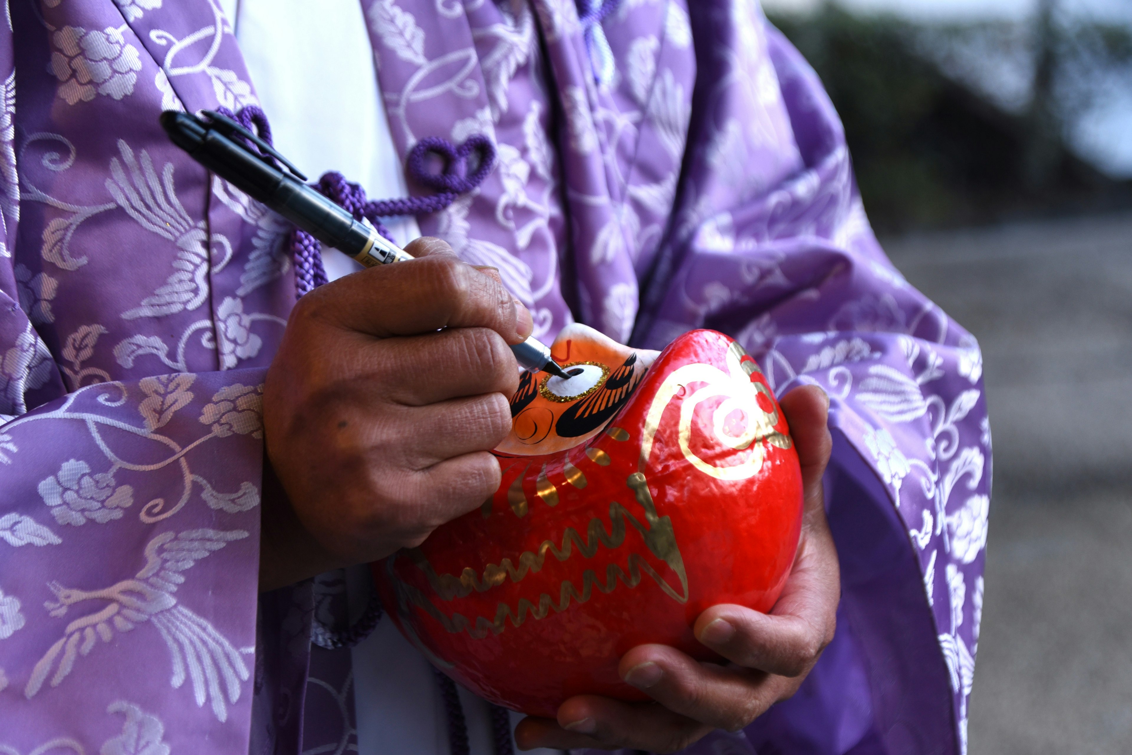 Person in lila Kleidung, die auf eine rote Daruma-Puppe malt