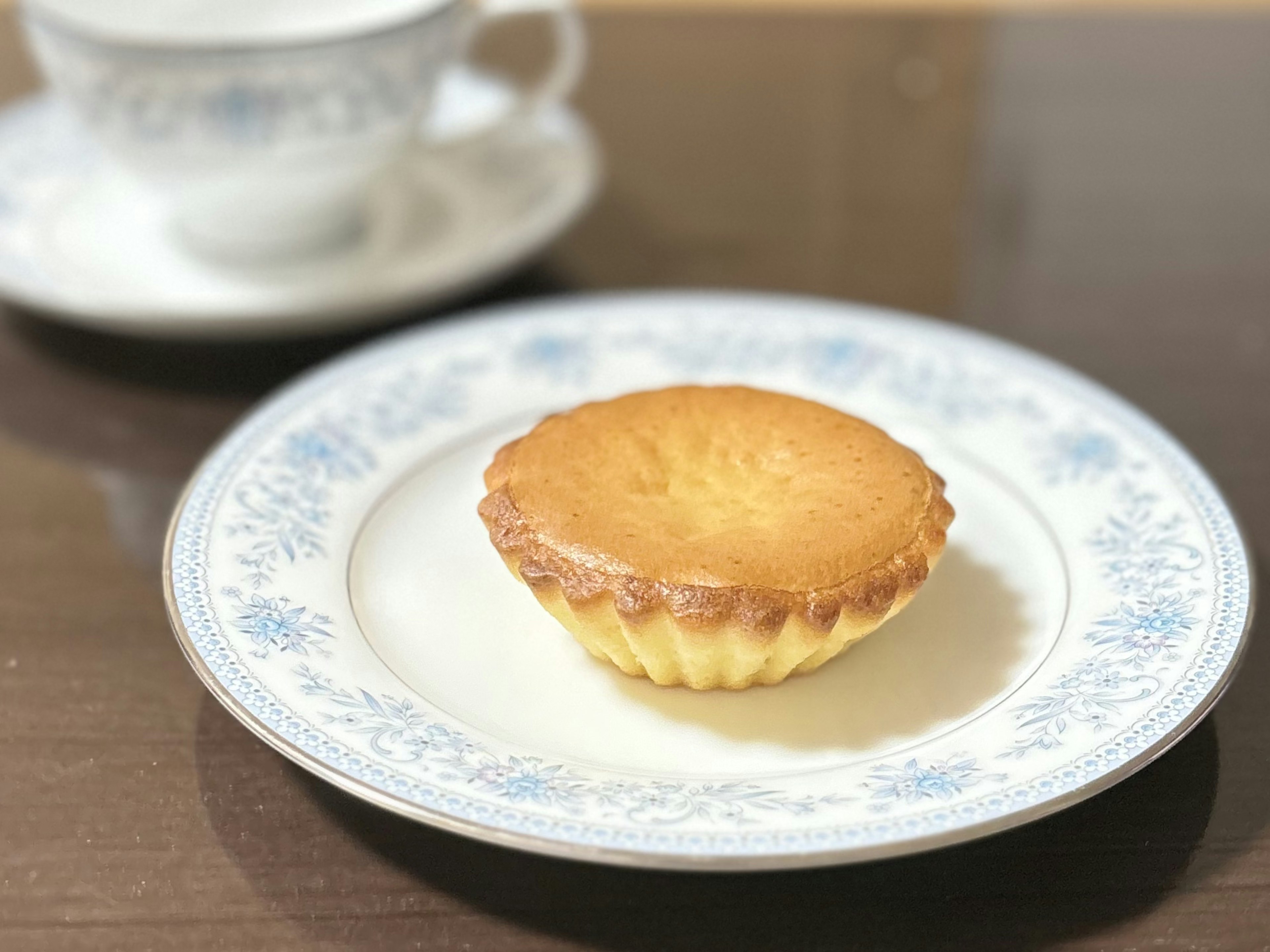 A golden brown cupcake on a blue floral plate with a cup in the background