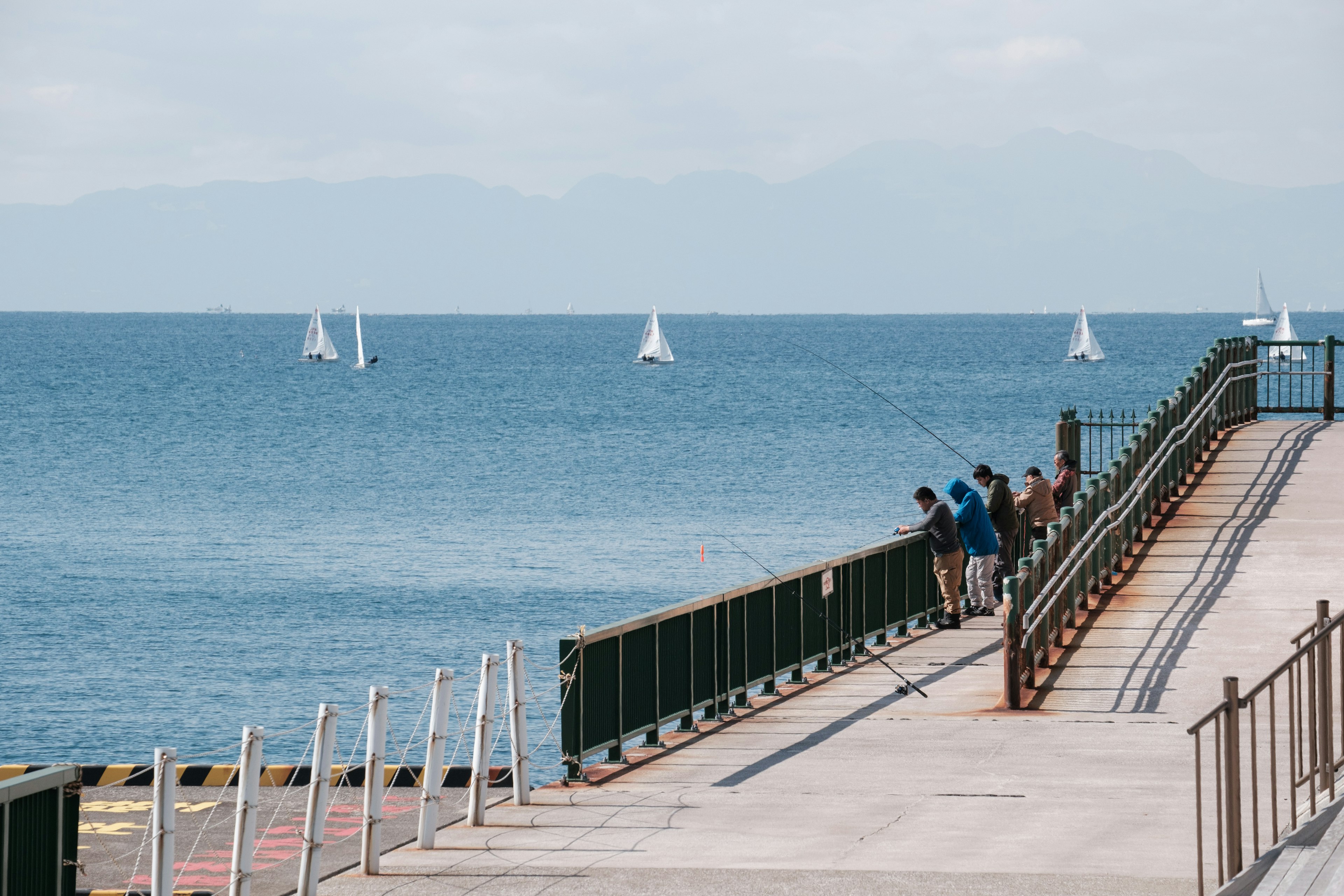 People standing on a seaside pier with sailboats in the background