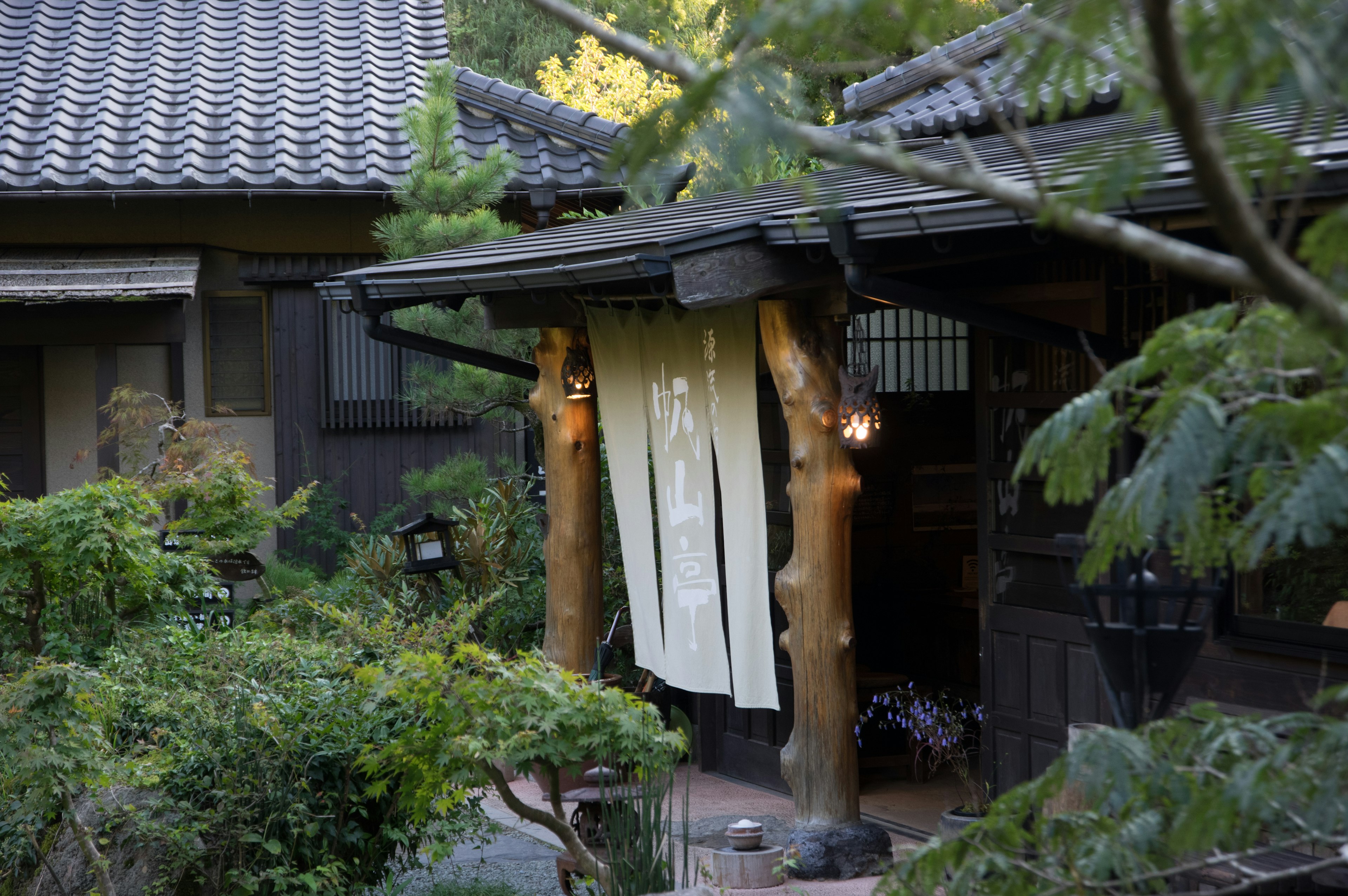 Entrada de una casa japonesa tradicional con un noren blanco y un jardín exuberante