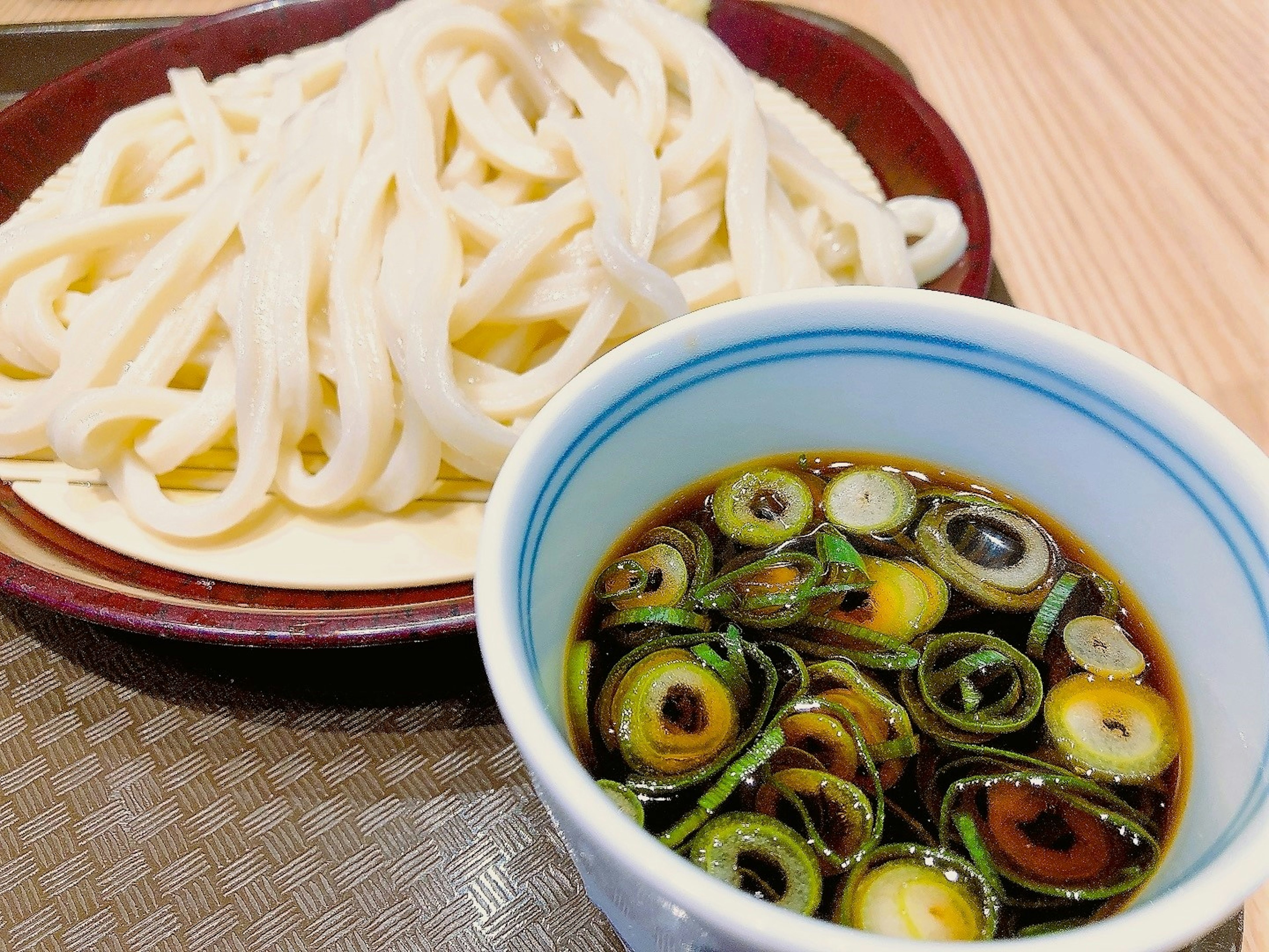 Sepiring mie udon disajikan dengan mangkuk saus celup yang dihiasi daun bawang