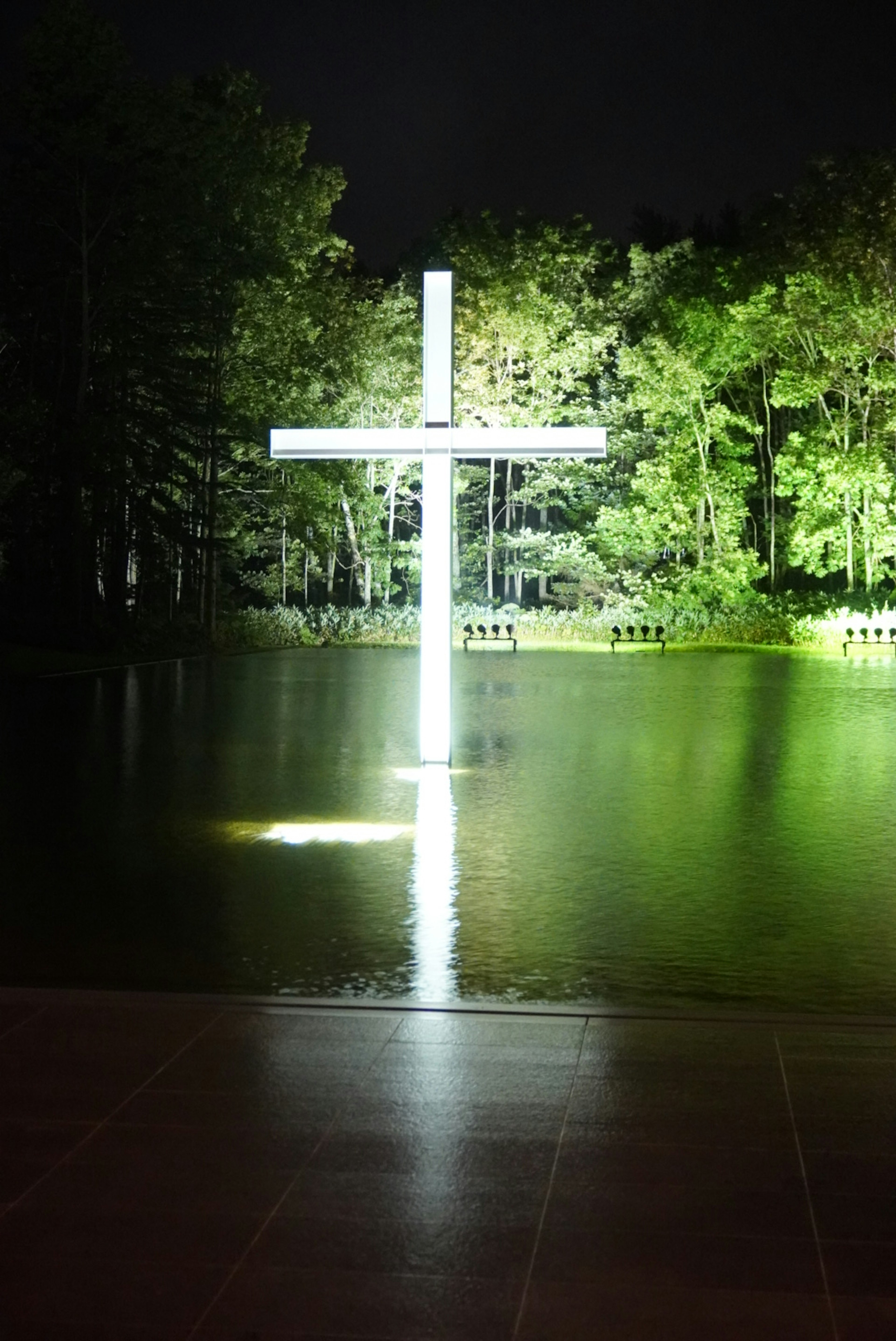 Ein beleuchtetes weißes Kreuz, das sich auf der Wasseroberfläche spiegelt, umgeben von grünen Bäumen bei Nacht